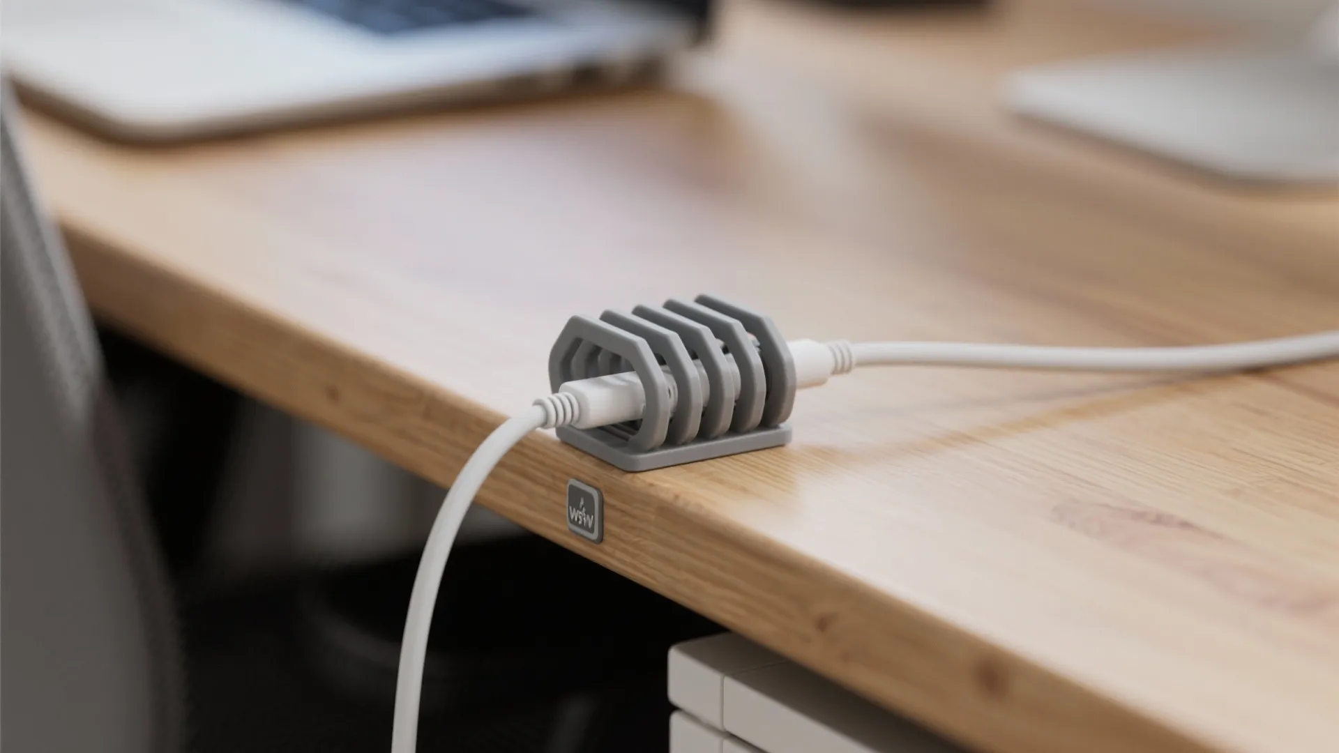 A small grey cable holder holds a white wire on the edge of a wooden desk