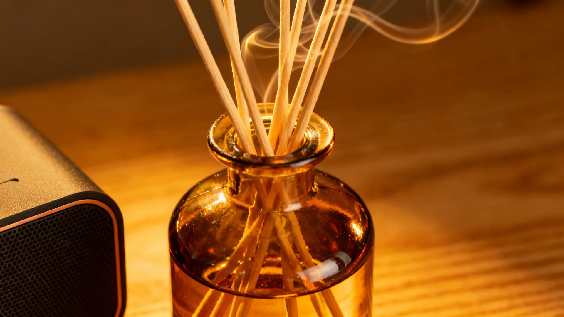 Amber glass diffuser with reeds glowing in warm light beside a compact speaker.