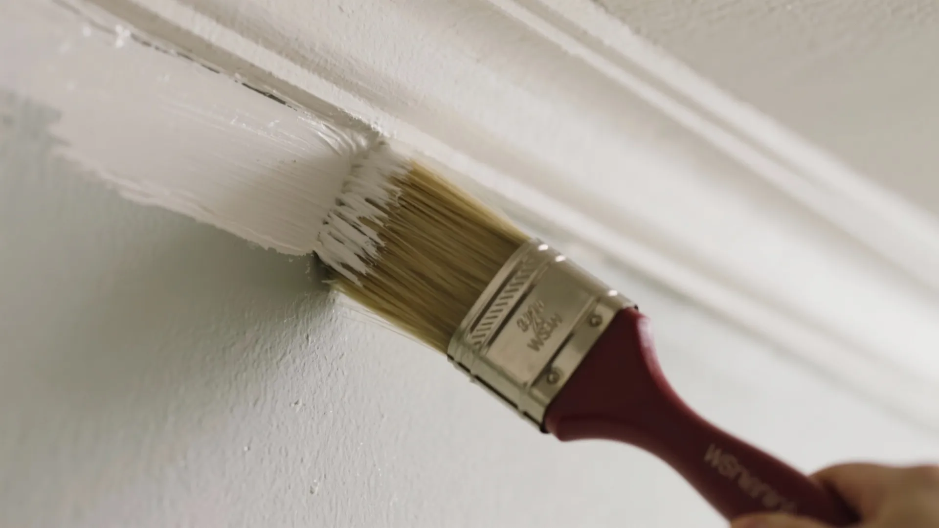 A hand using a paint brush to apply white paint along the edge of wall