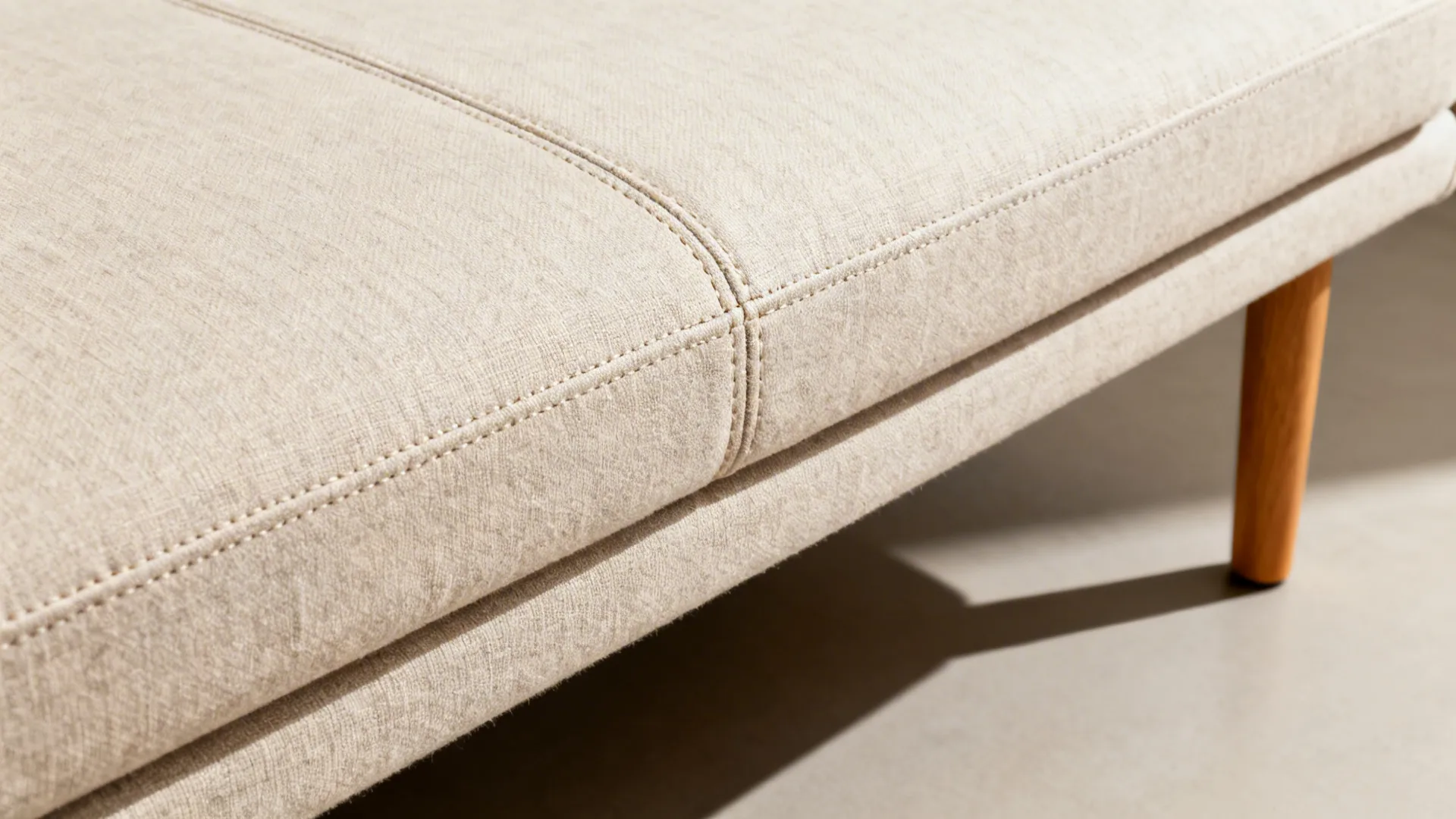 Close up of beige fabric sofa with neat stitching and a light brown wooden leg