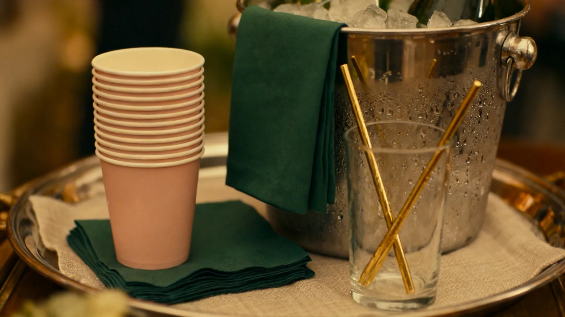 Close-up of blush cups, green napkins, and gold stirrers beside a dewy ice bucket on a tray.