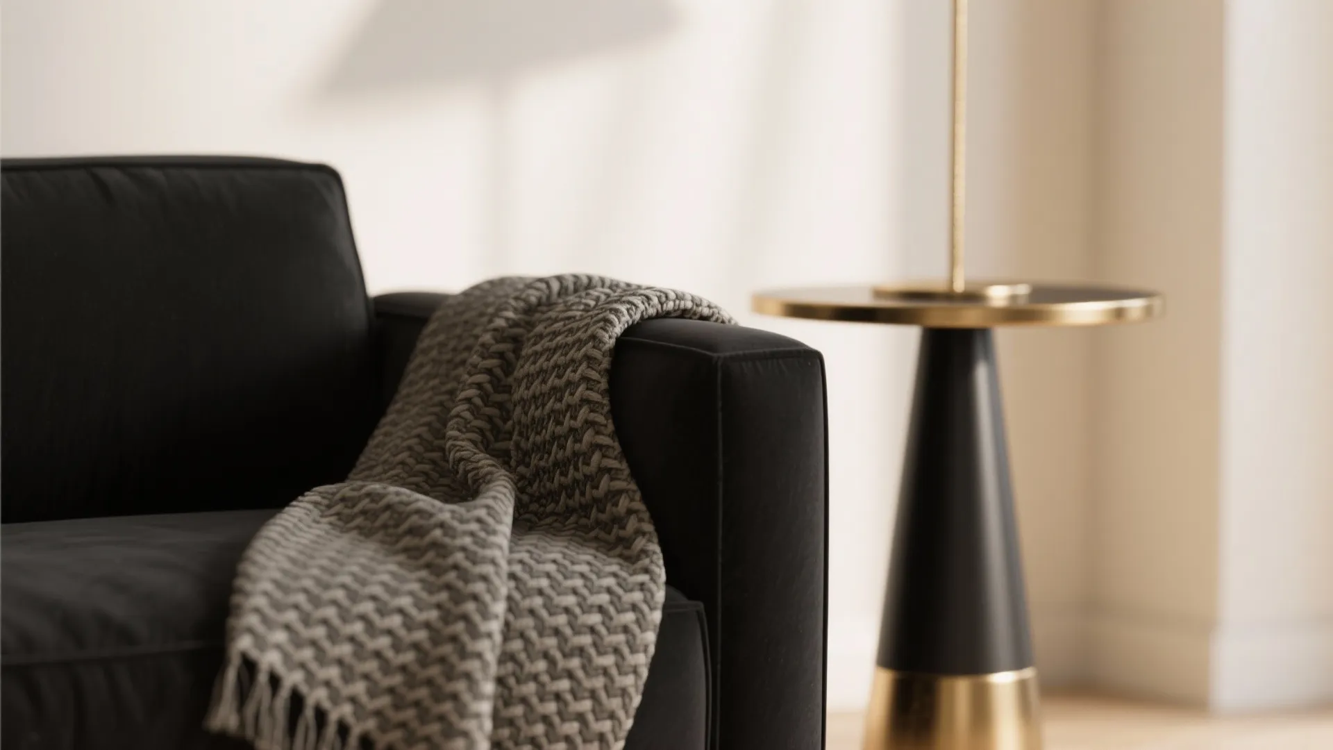 Close-up of a matte black sofa with a woven throw and mixed-metal side table against a warm white wall.