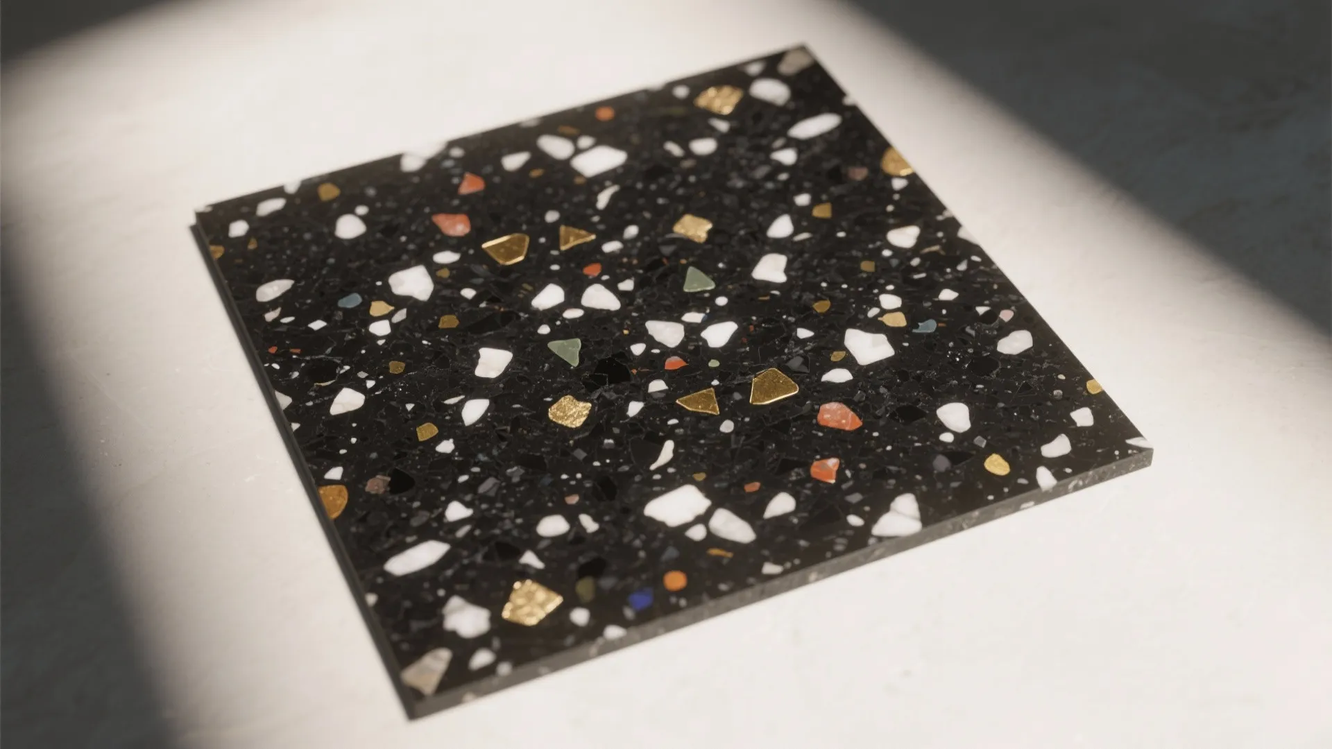 A square sample of black terrazzo tile with colorful stone chips on a white surface