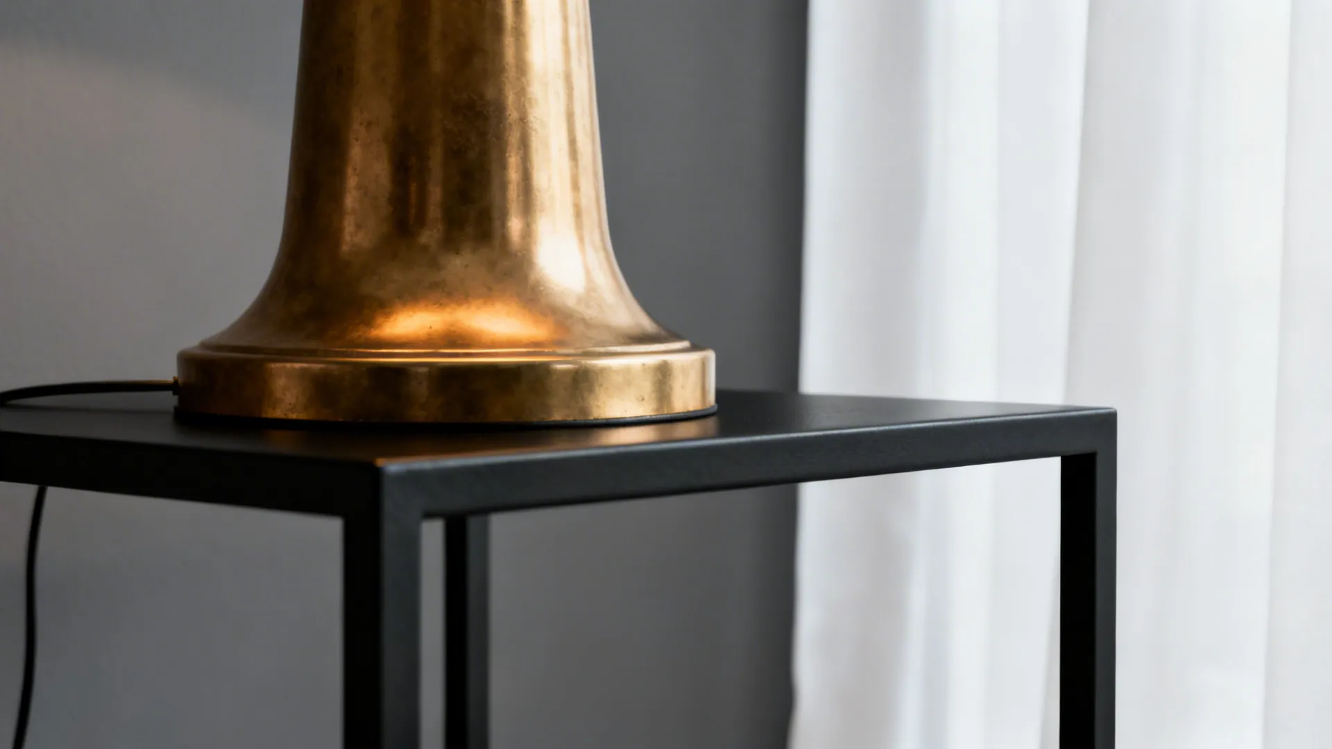 Gold lamp base sitting on a black metal table against a grey wall and white curtains