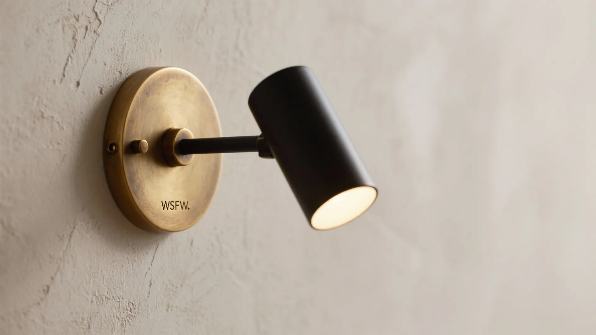 Small black adjustable wall light with brass base mounted on a textured white plaster wall