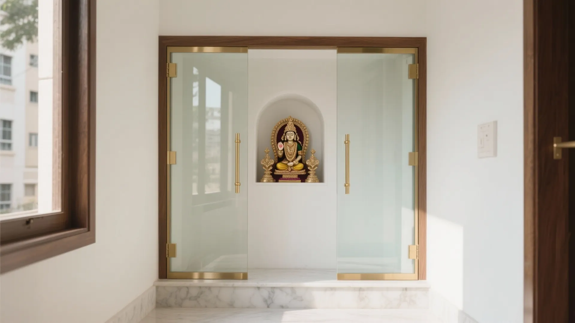 Minimalist mandir niche with clear tempered double glass doors and slim satin brass pulls in a bright compact foyer.