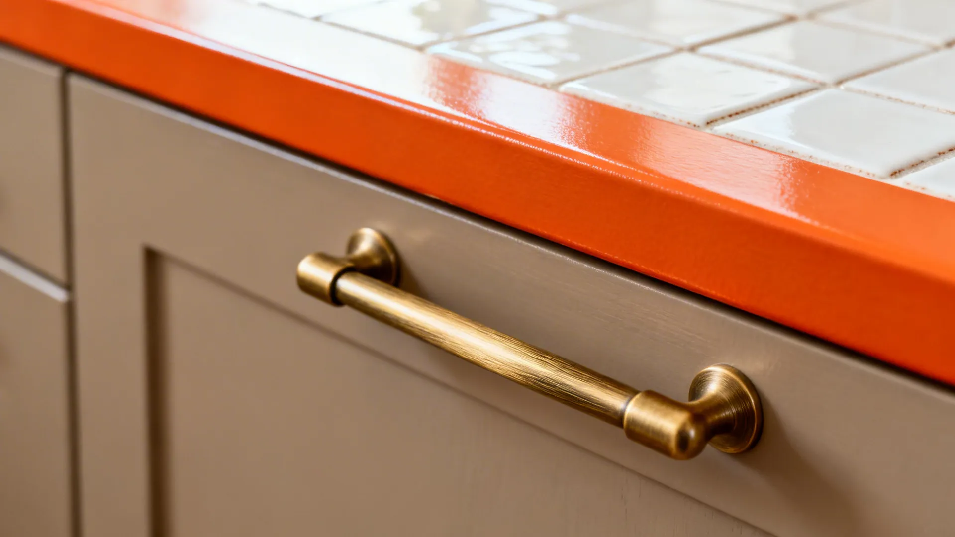 Macro detail of brushed brass pull on greige cabinet beside orange countertop and glazed tile.