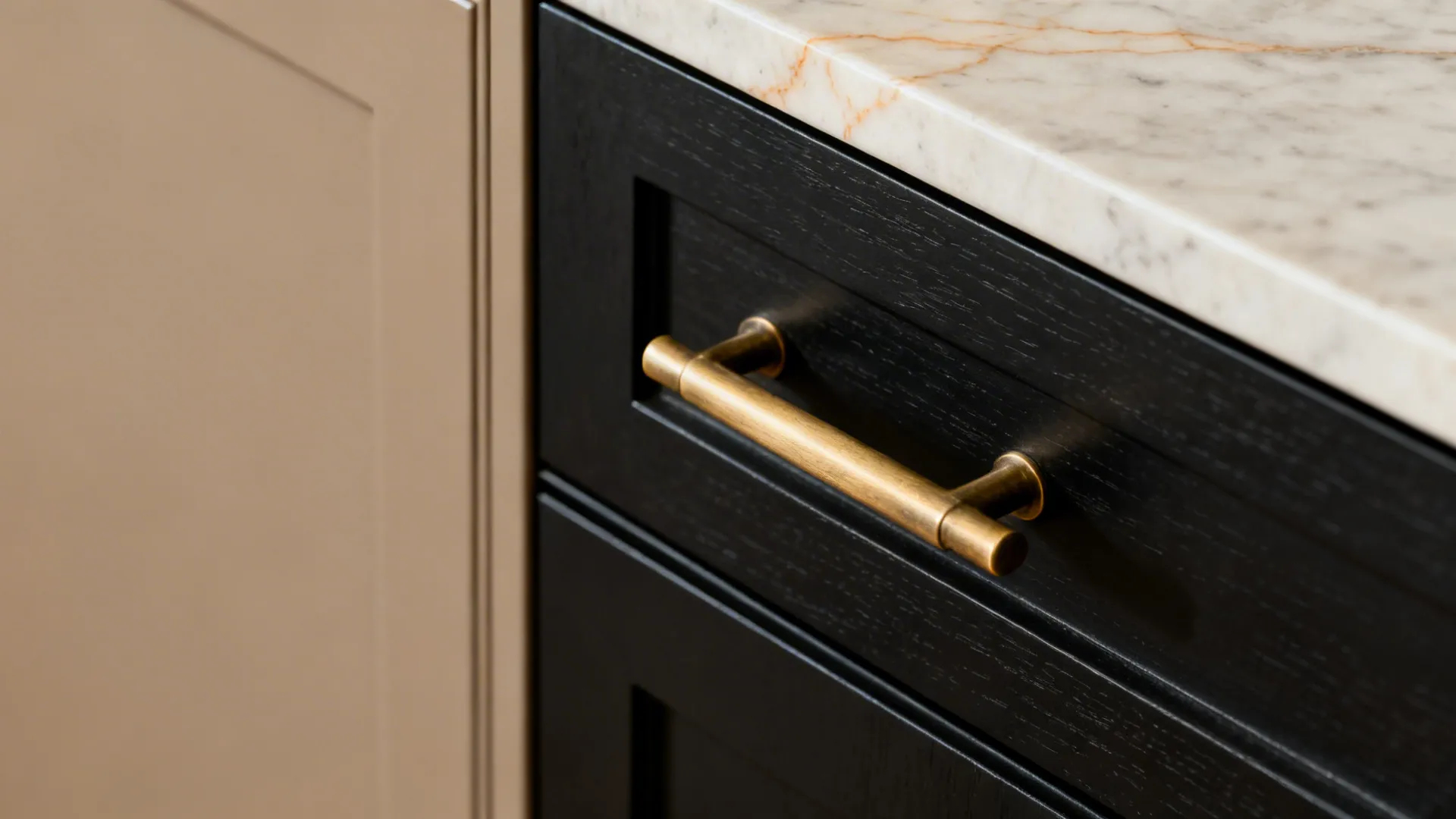 Macro of brushed brass pull on matte black cabinet beside warm greige door and quartz top.
