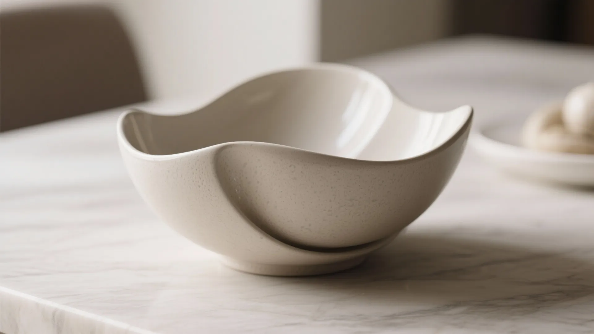 Modern cream colored ceramic bowl with curved edges placed on a white marble dining table