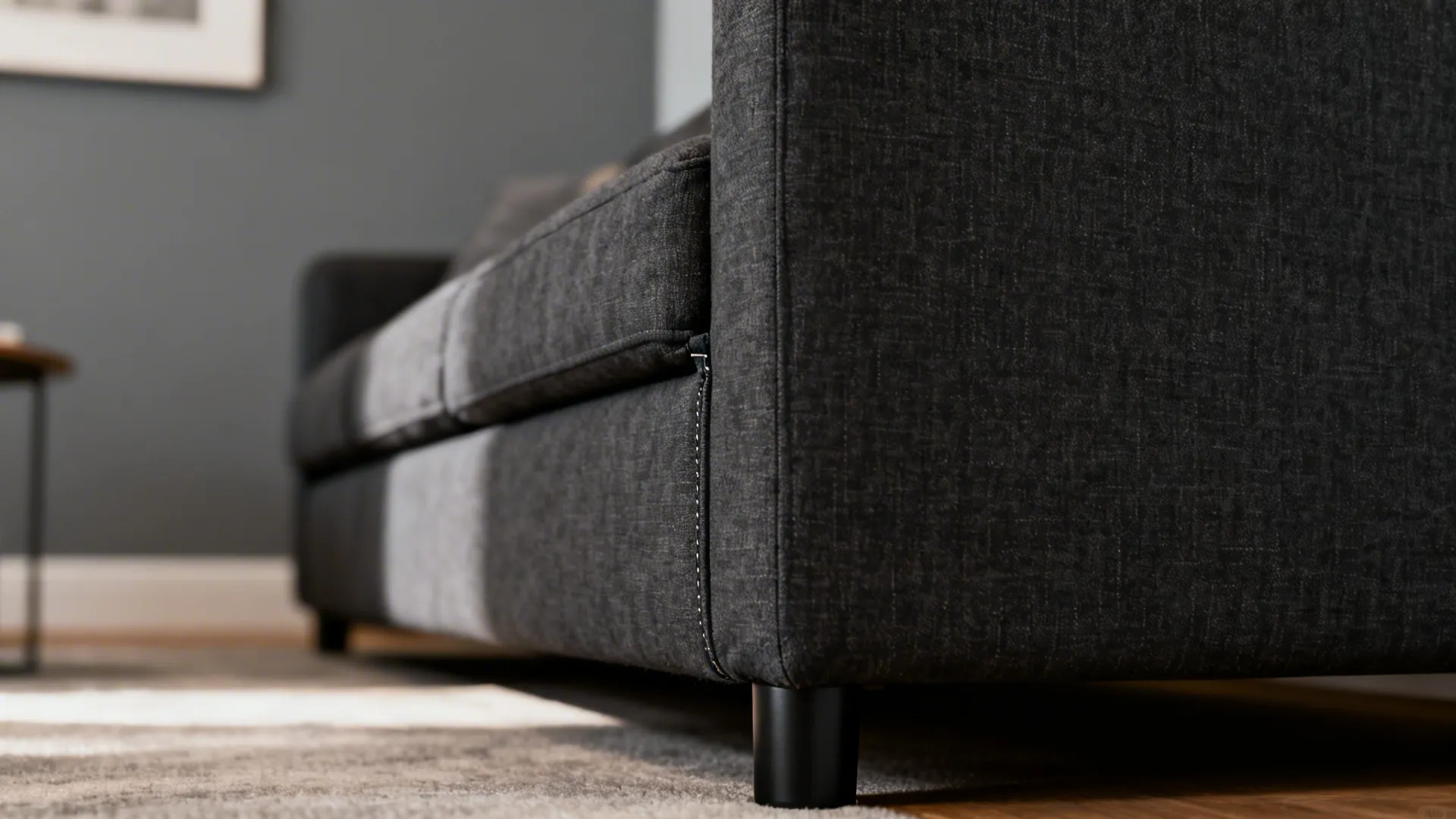 Close-up of charcoal grey sectional fabric, matte black leg and mid-tone grey wall.