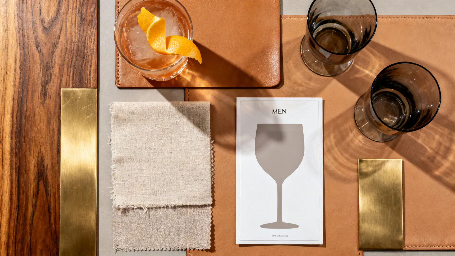 Flatlay of timber, brass, tan leather, linen, and smoky glass for a dining mood board.