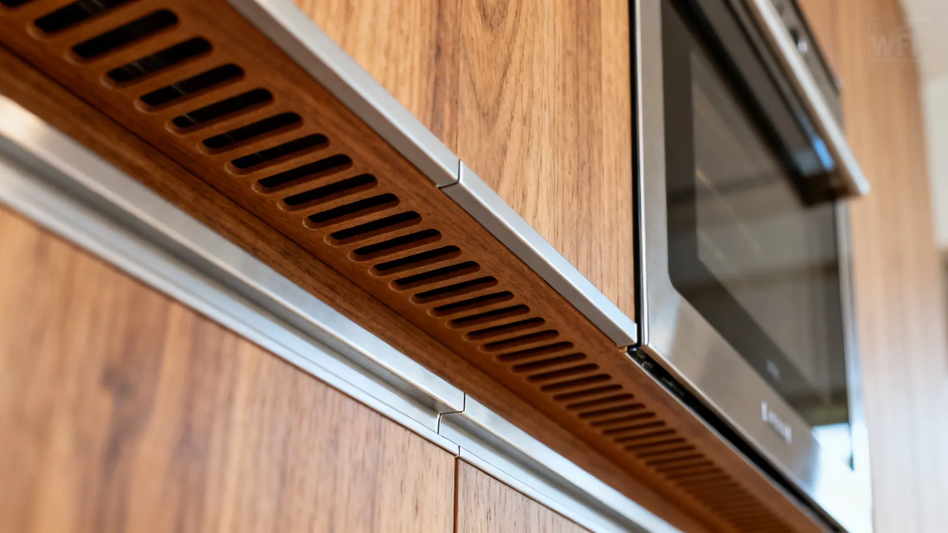 Close-up of concealed vent slots under a wood panel near a built-in oven with protective edging.