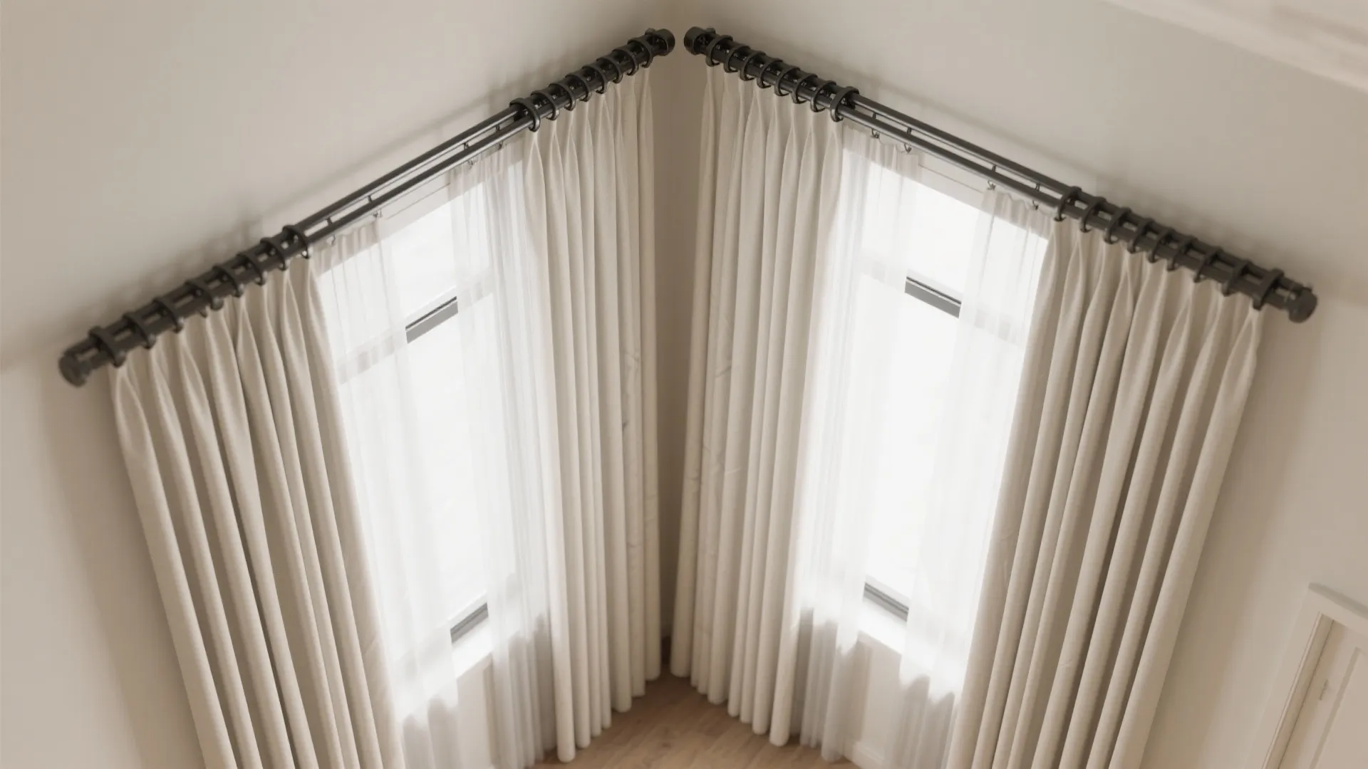 Corner windows with cream curtains on black metal rods in a room with light wooden floors