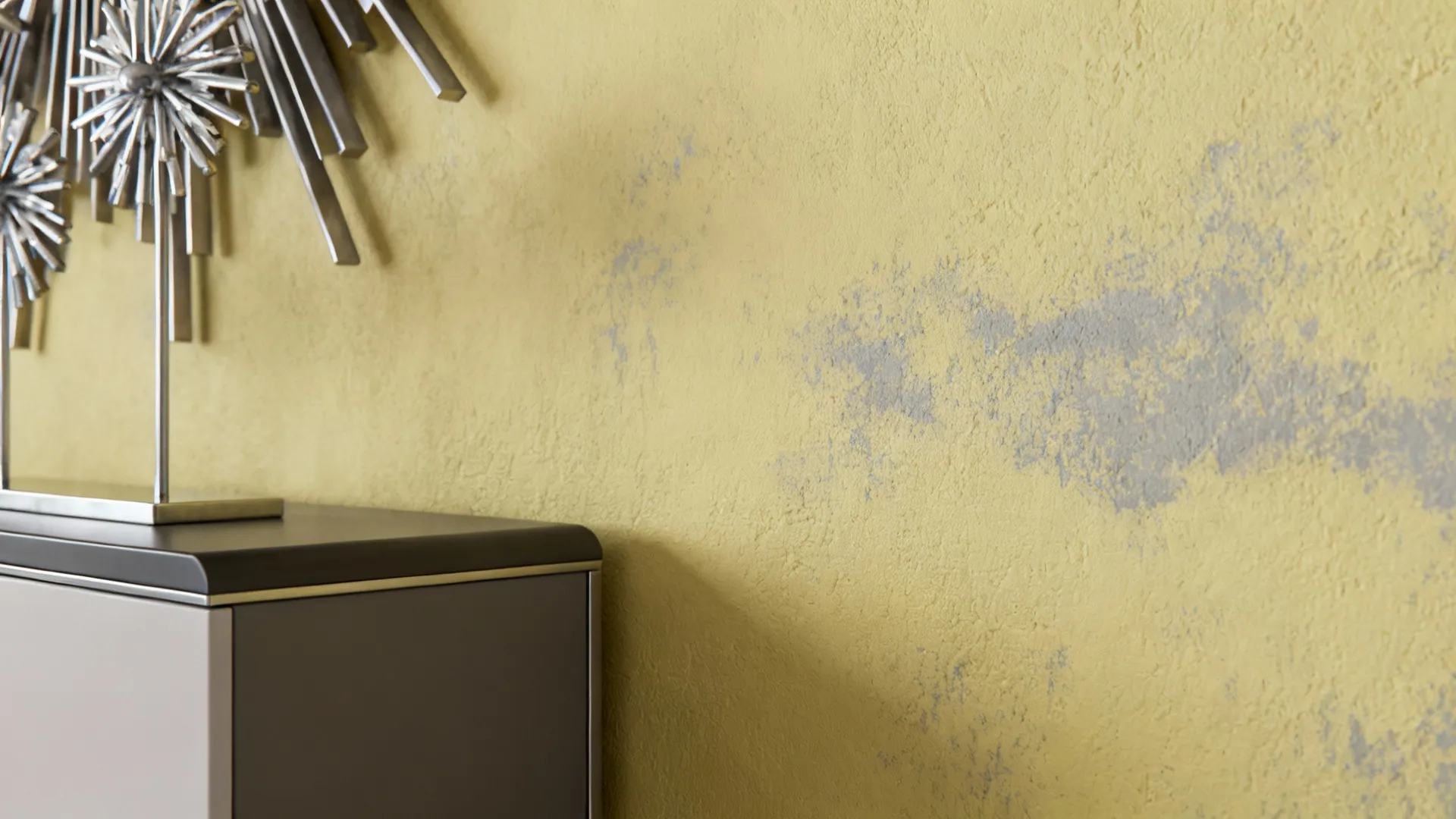 Close-up of creamy yellow wall with subtle gray undertone beside cool metal decor showing sophisticated muted tones.