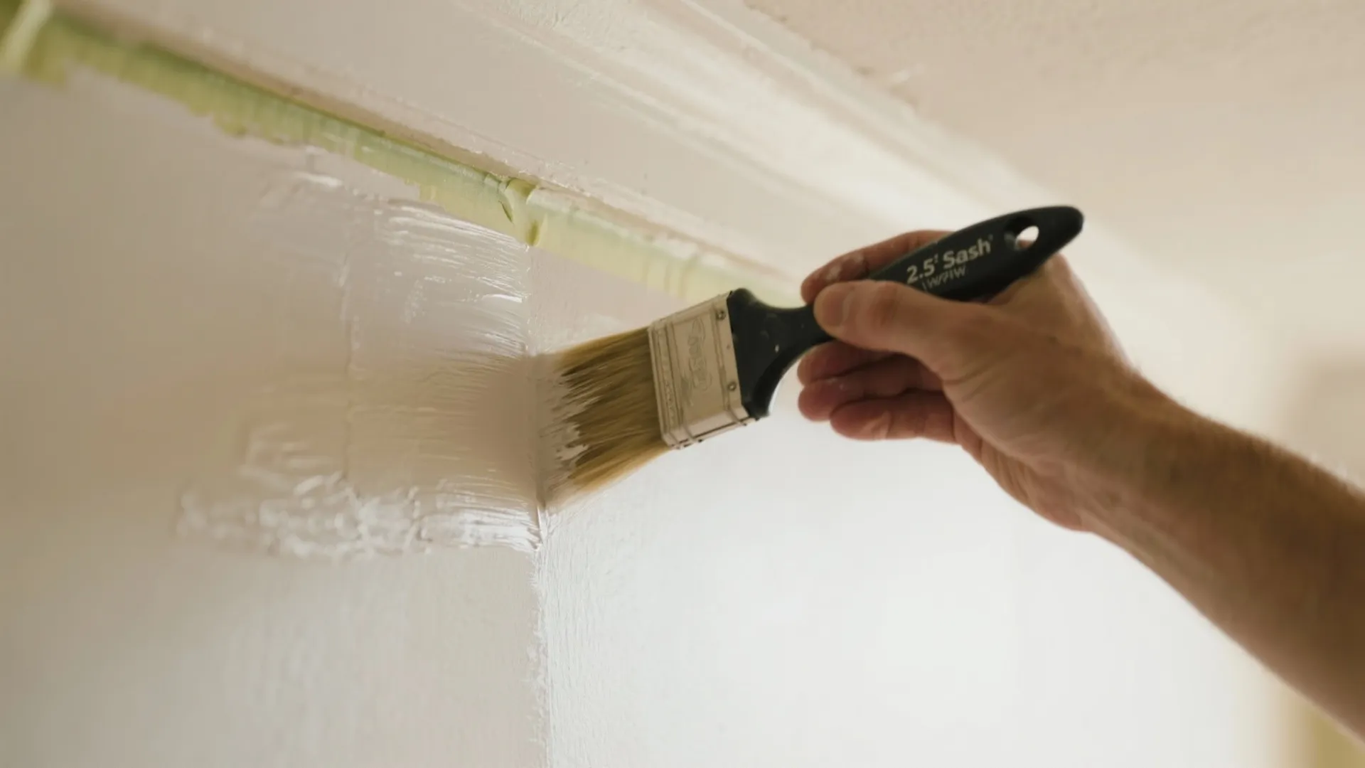 Hand using a black paint brush to apply white paint on a wall with tape