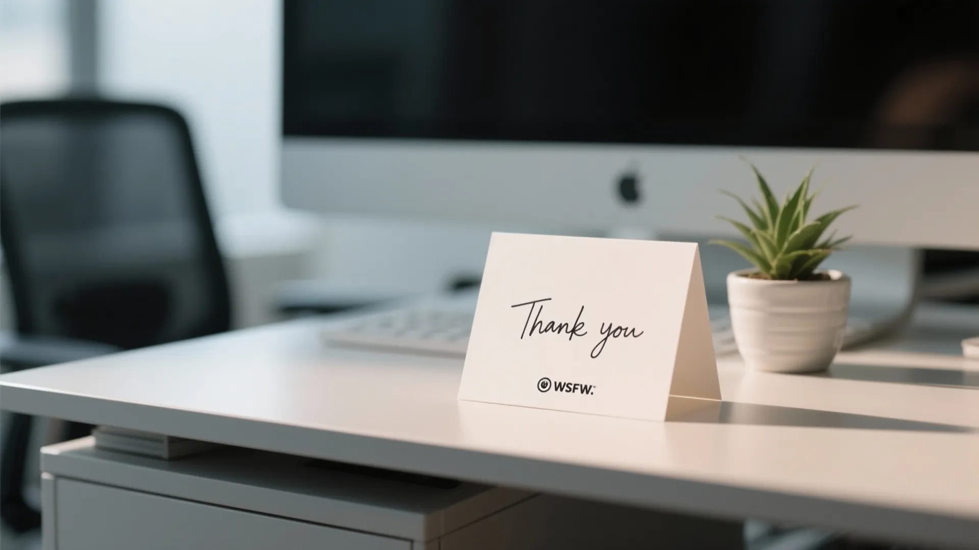 White thank you card on a white desk with a computer monitor and small plant