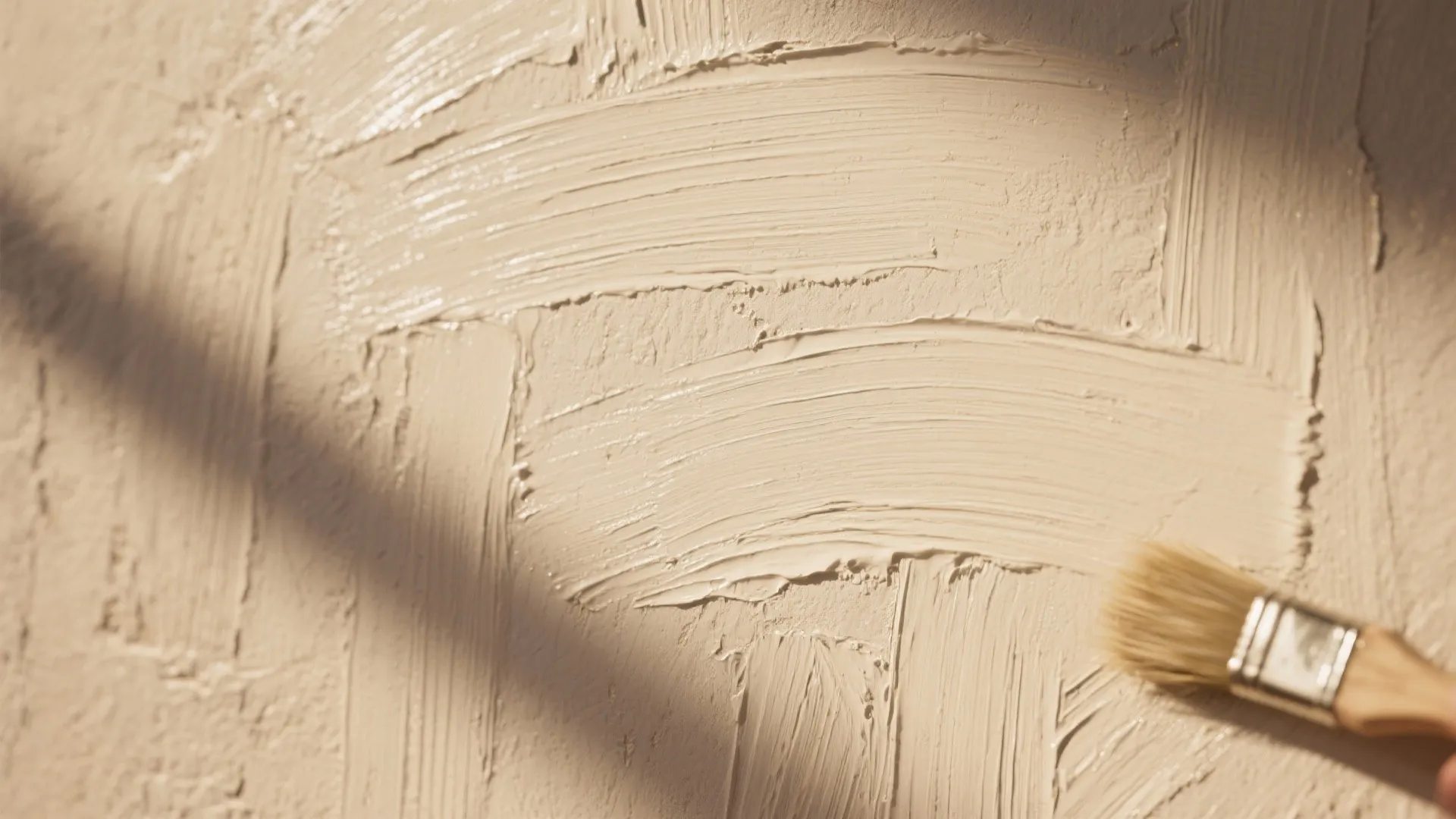 A small paint brush applying beige paint with thick texture onto a flat wall surface