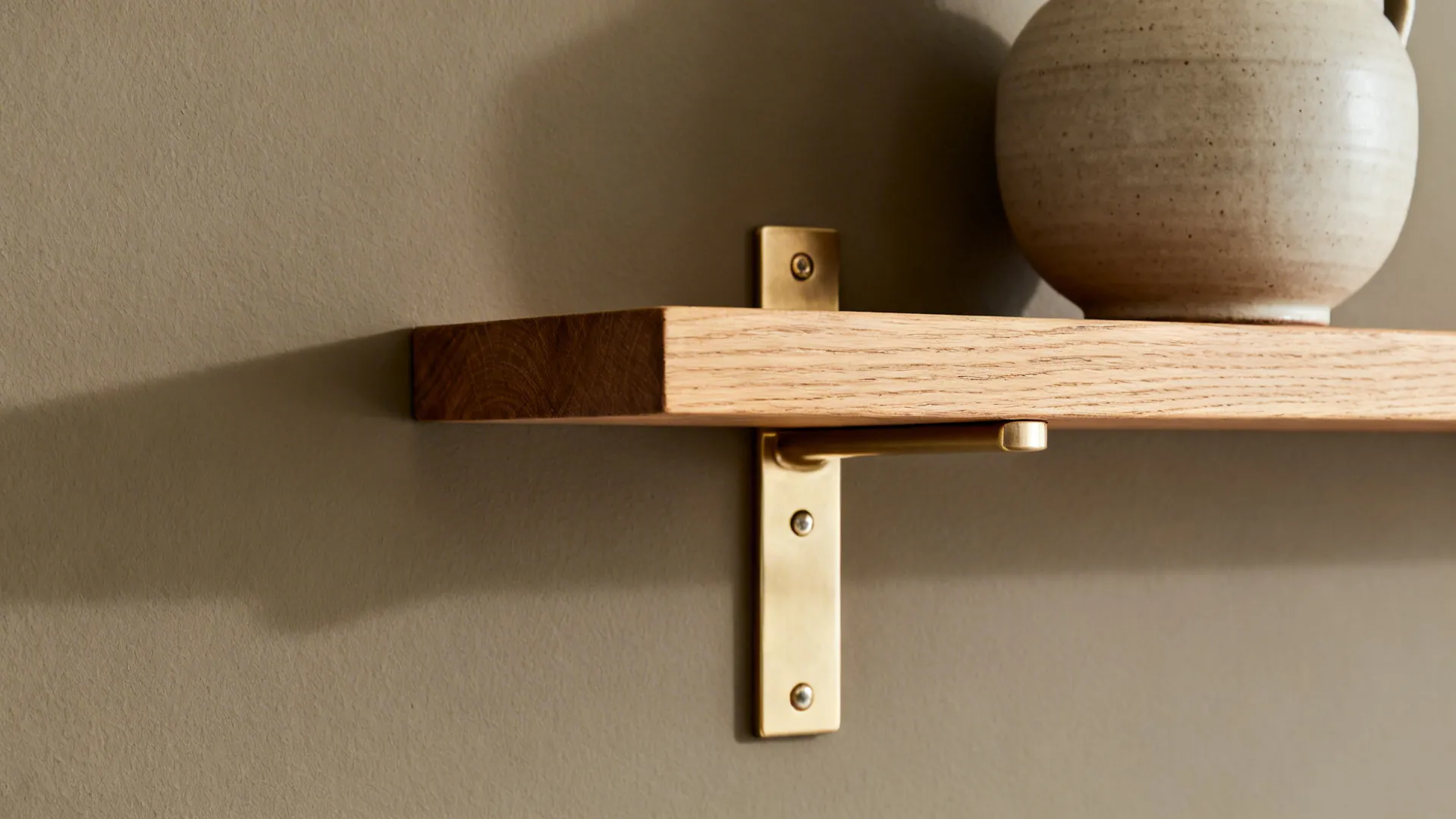 Close-up of clay-gray wall with oak shelf and brushed metal hardware in soft daylight.