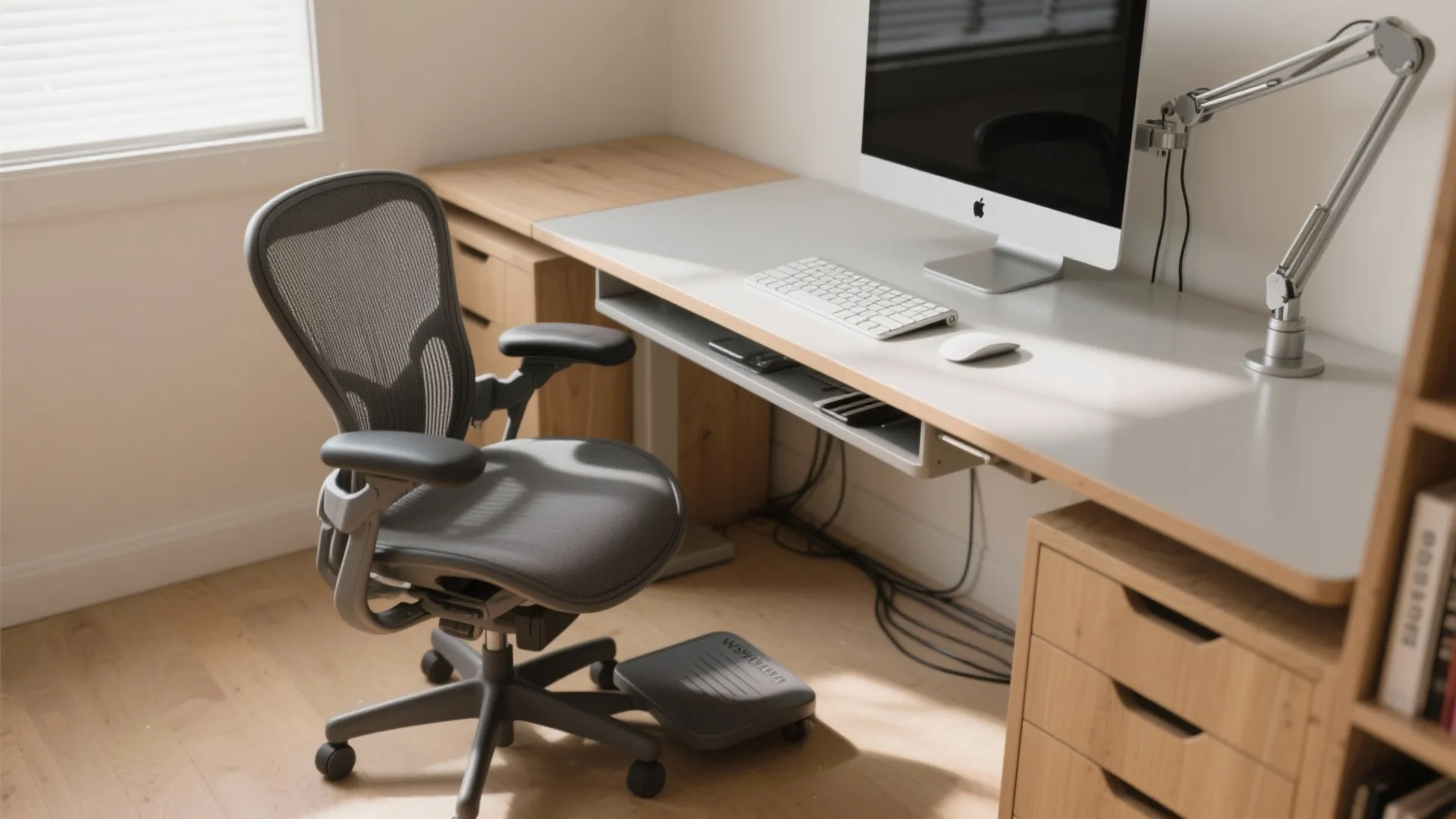 Ergonomic grey office chair with footrest at a wooden desk with computer and desk light