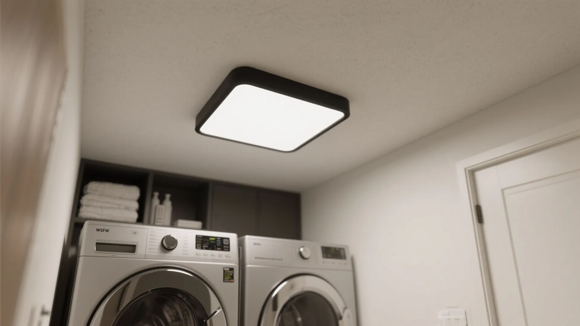 Square black ceiling light fixture mounted on white ceiling above silver washing machines in laundry space