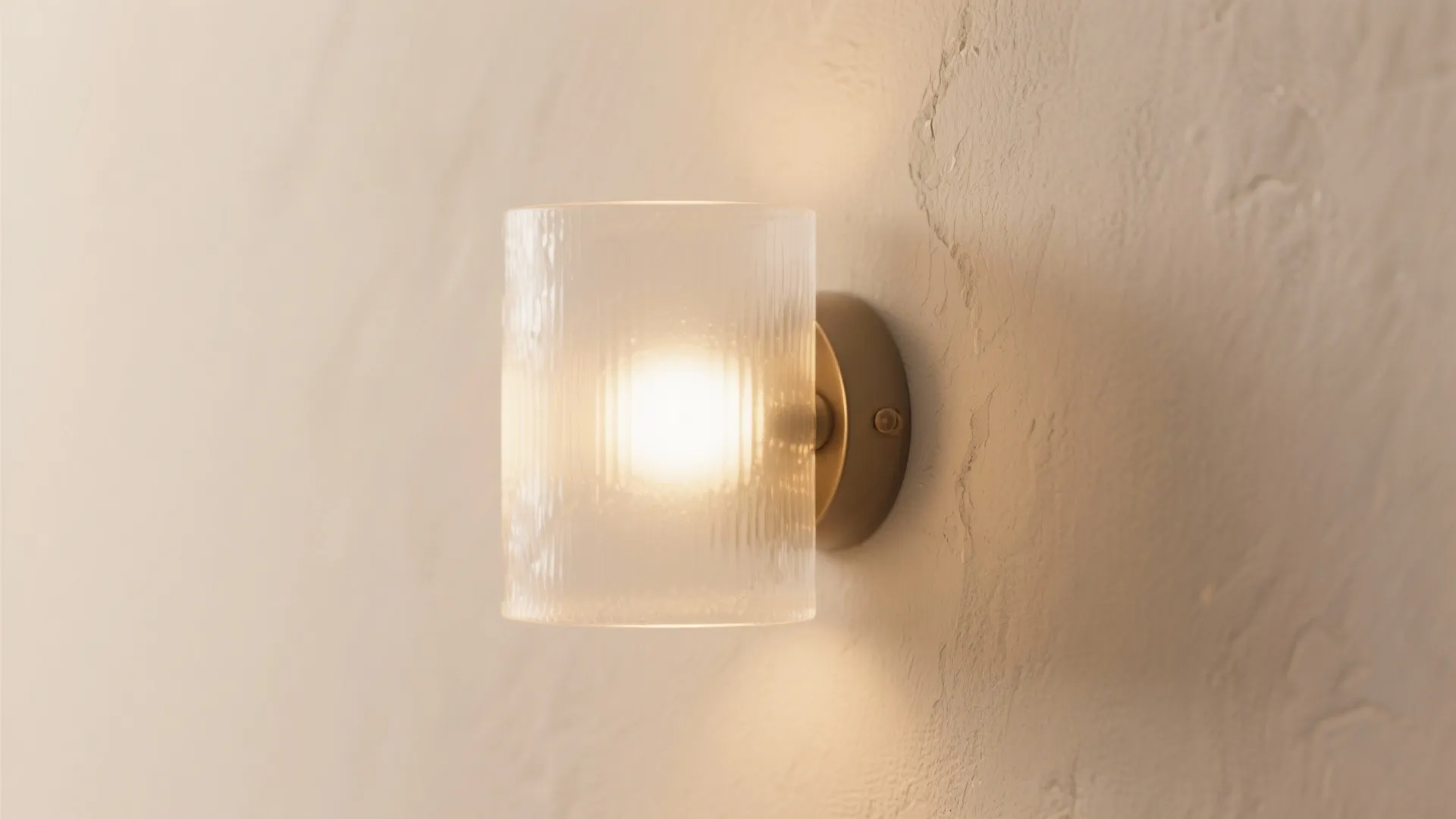 A modern cylindrical glass wall light glowing with warm yellow light on a white wall