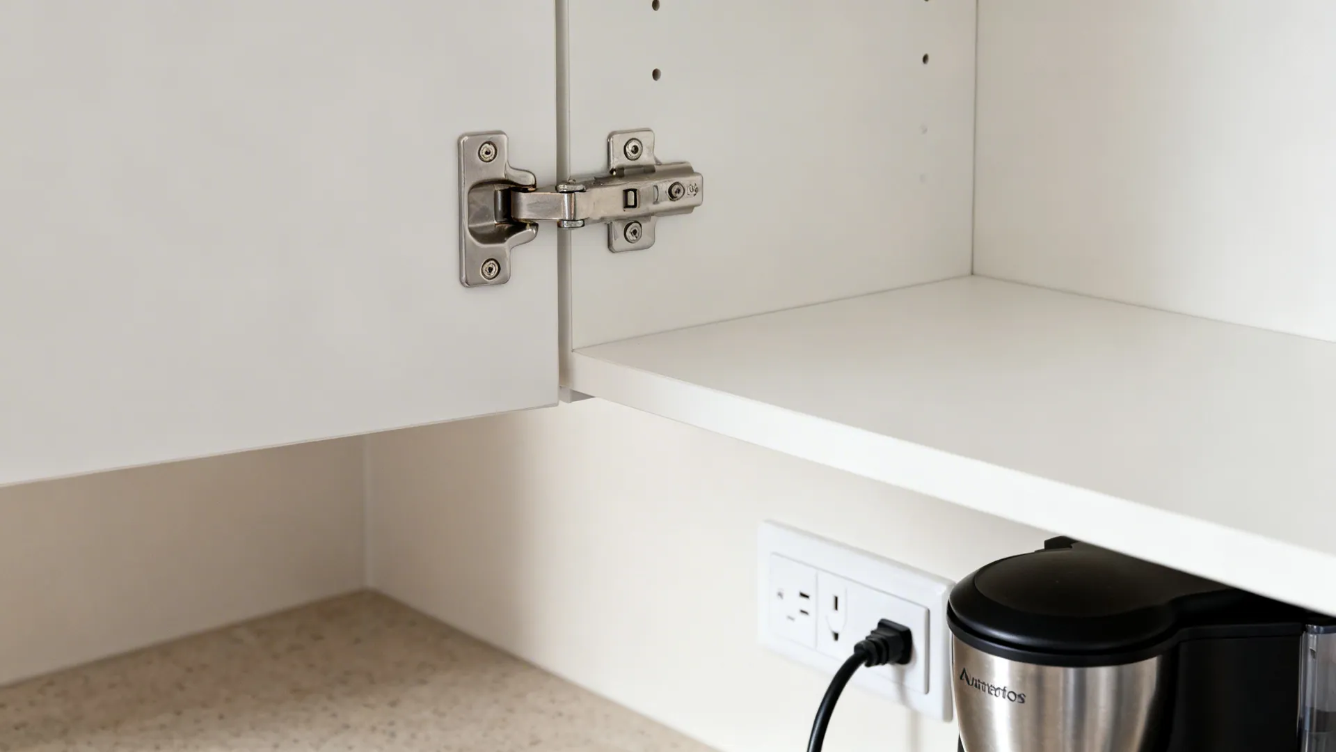 Macro of flip-up cabinet hardware and a discreet in-cabinet outlet beside an espresso machine.