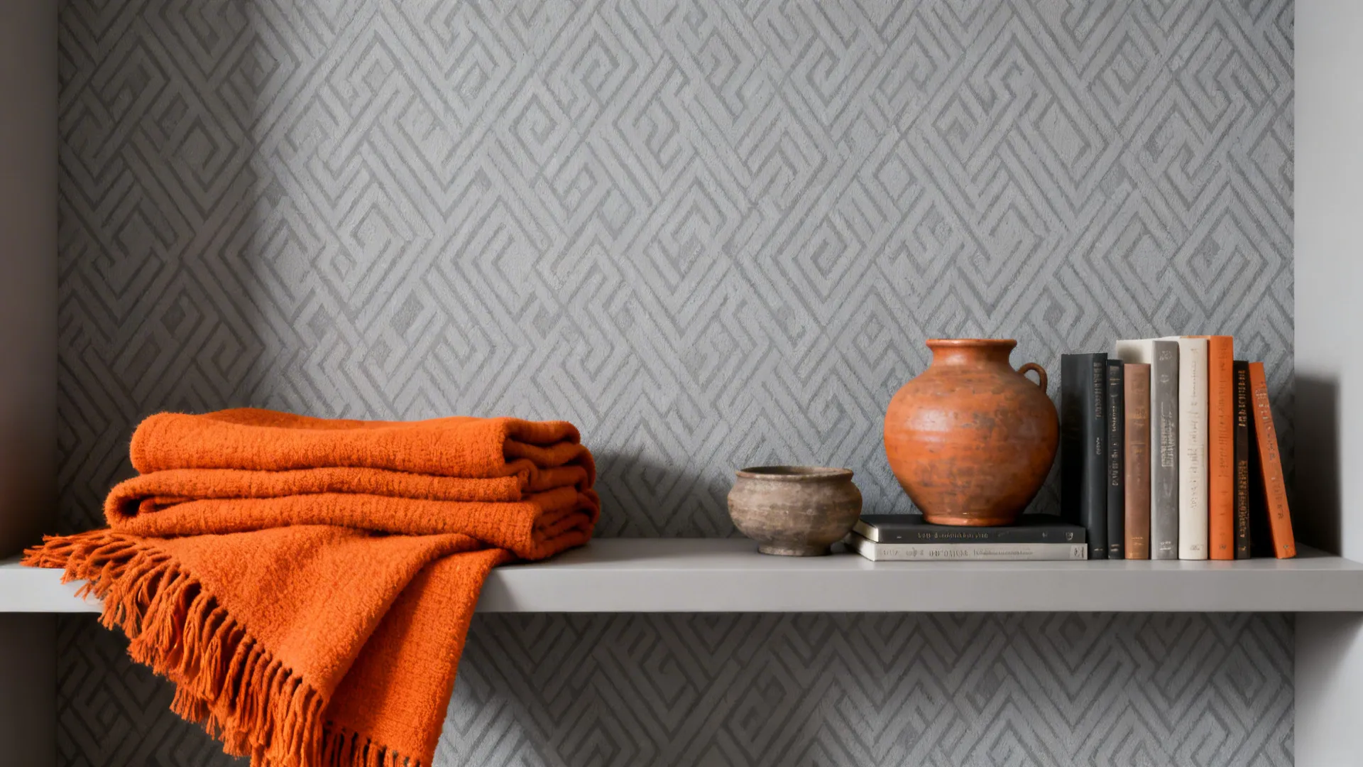 Subtle geometric gray wallpaper with orange throws and pottery arranged on a shelf.