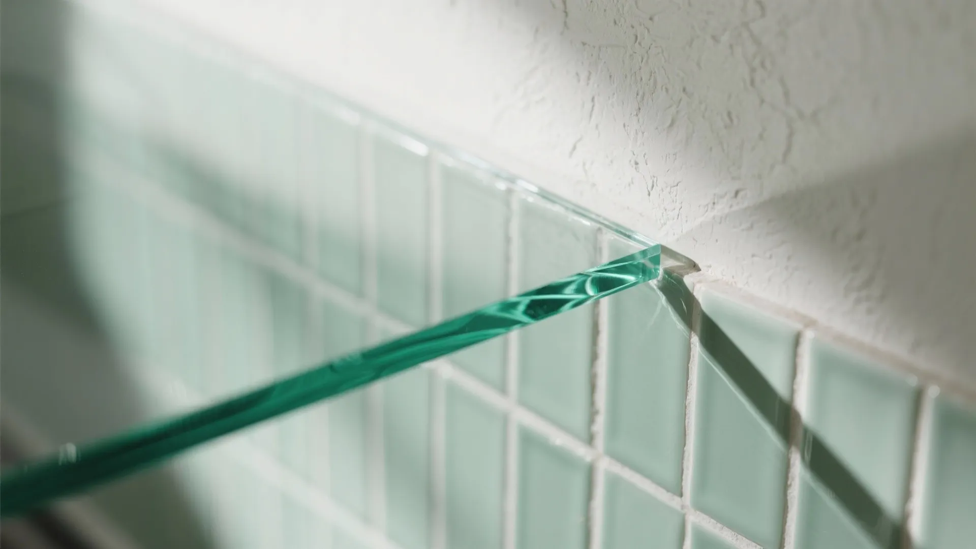 Close up of glass shelf edge fixed against light green wall tiles and plaster wall