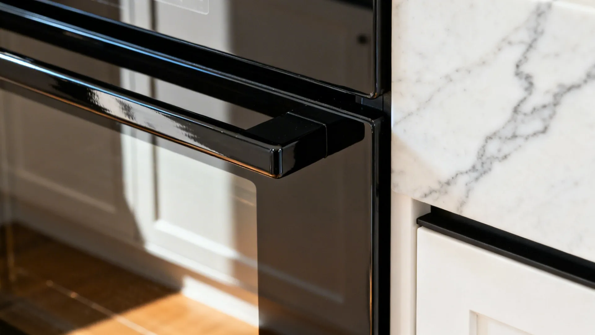 Glossy black oven edge reflecting warm white cabinet and slim black pull on quartz.