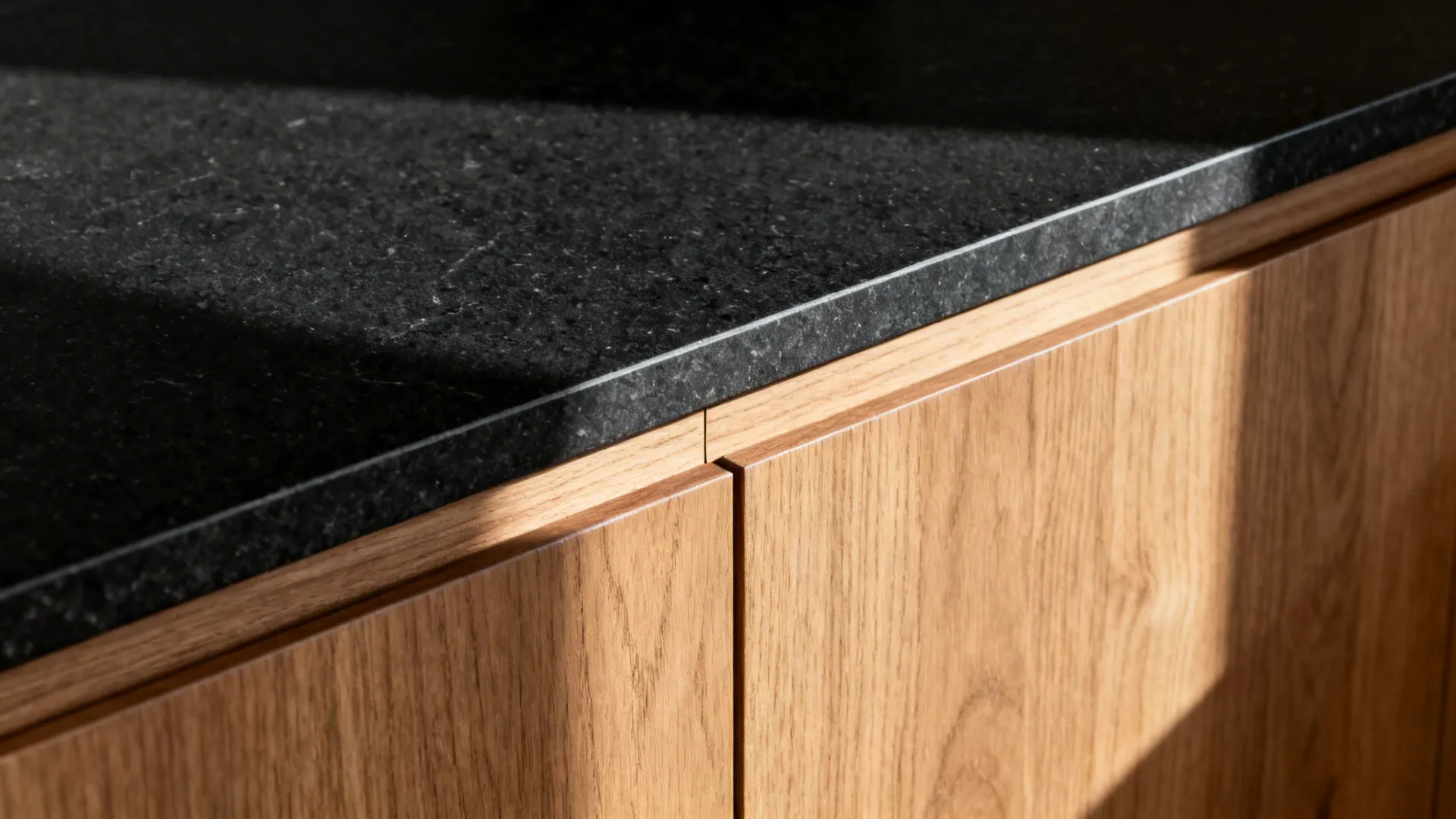 Macro of honed black granite edge and rift-cut oak handleless cabinet groove.