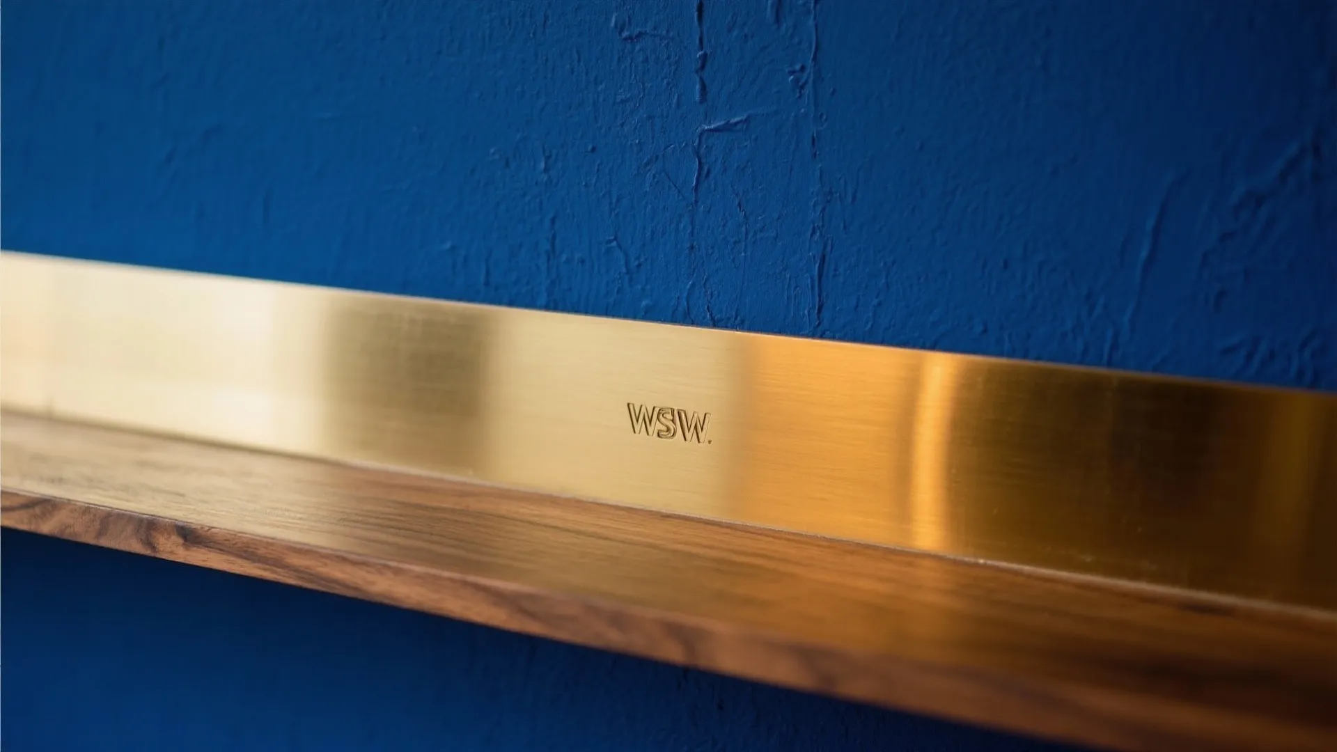 Macro of matte indigo wall with warm light reflecting on brass beside a wood shelf.