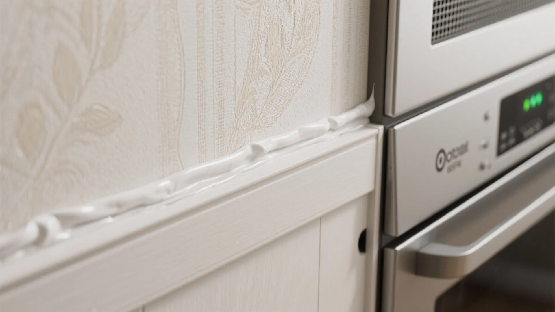 Close-up of wallpaper edge sealed with caulk near an appliance gap and ventilation details.