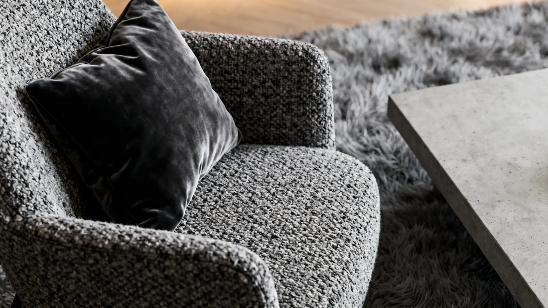 Close-up of velvet cushions, boucle chair, grey rug and concrete coffee table edge.
