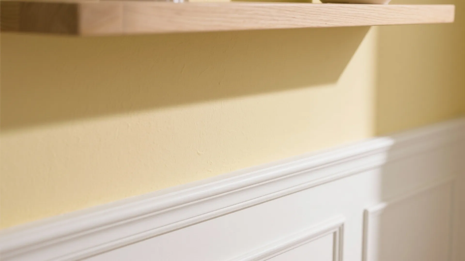 Close up of light yellow wall with white wall panel and wooden shelf in bright room