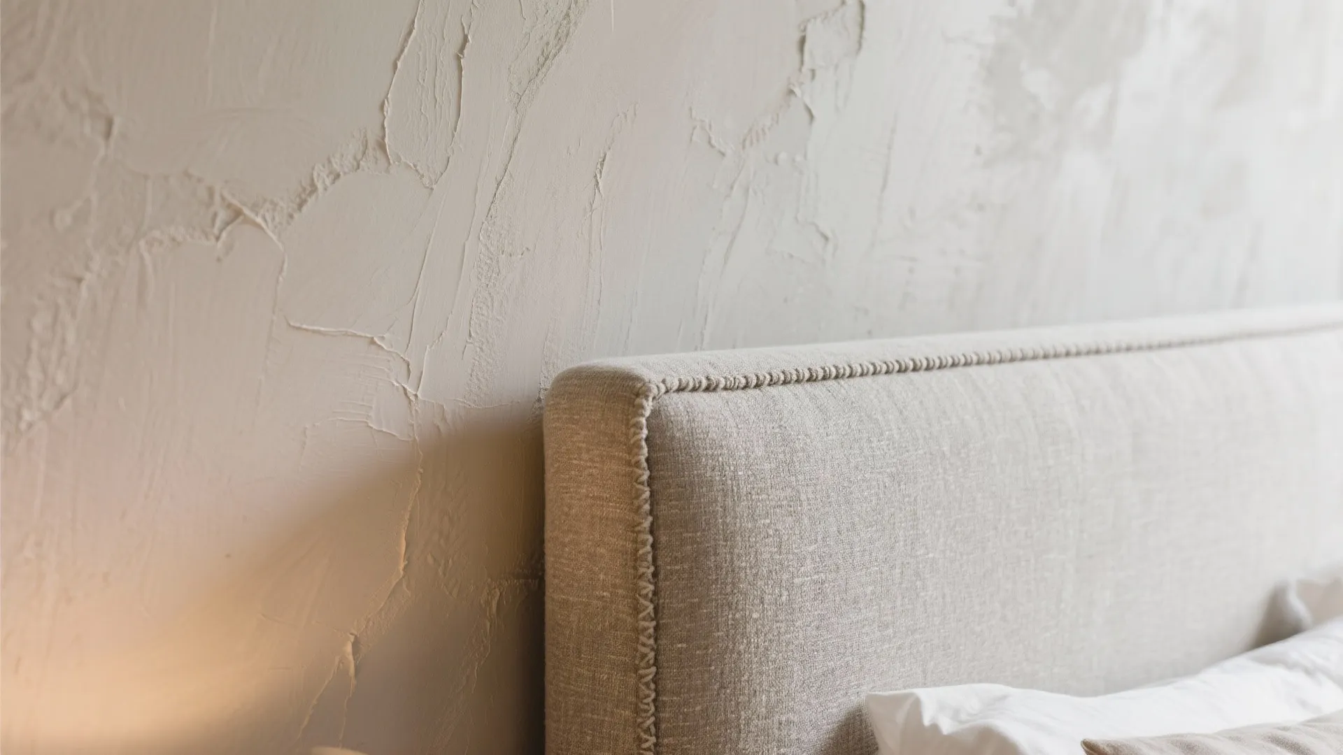 Close up of a beige fabric headboard against a textured light grey plaster bedroom wall surface