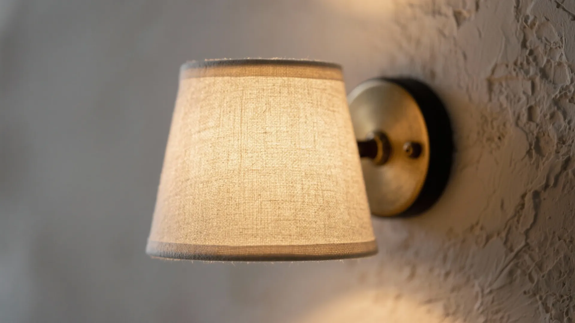Glowing wall light with a beige fabric shade mounted on a white textured interior plaster wall