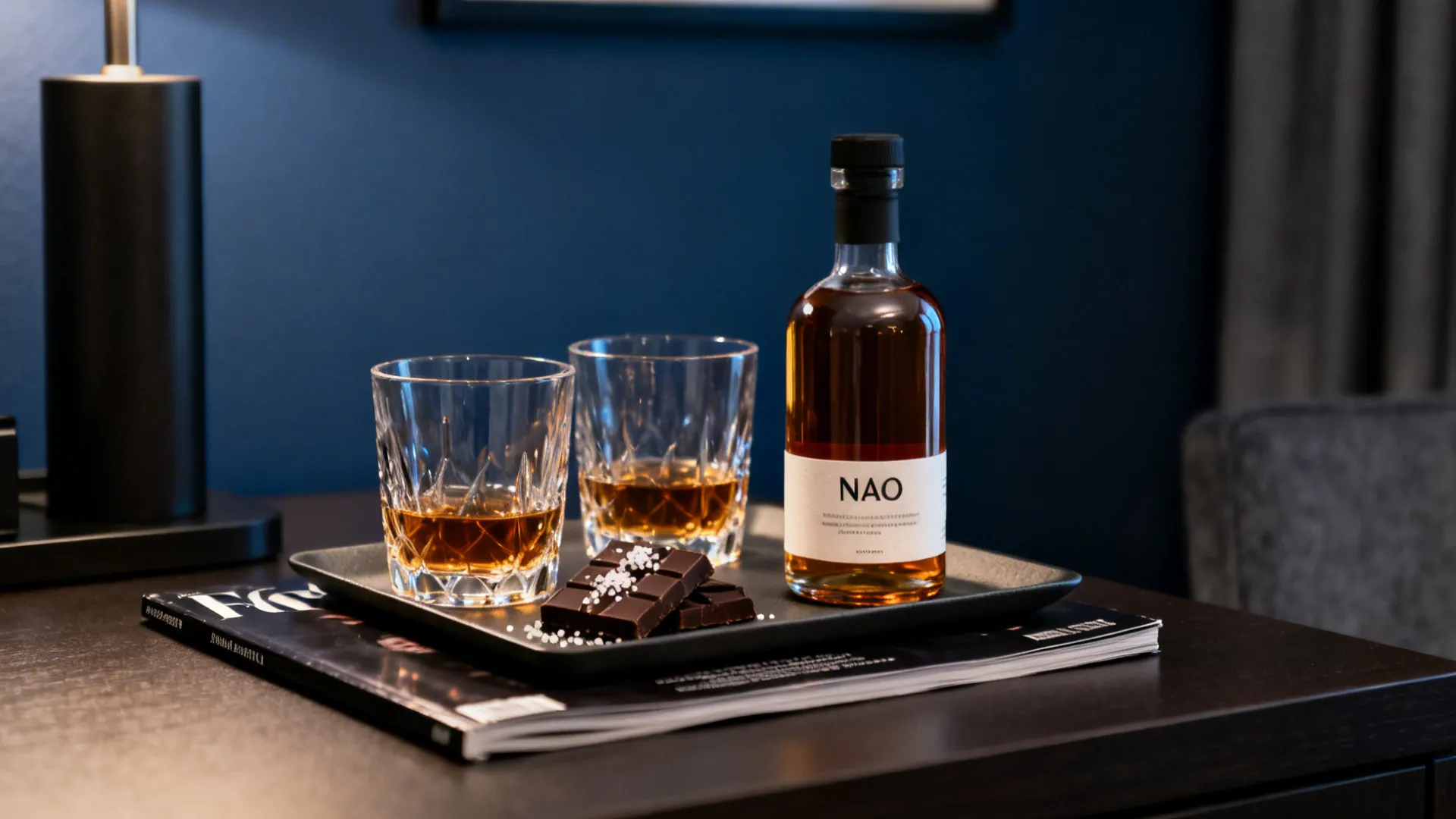 Two rocks glasses, an unbranded bottle, and dark chocolate arranged neatly on a hotel desk.