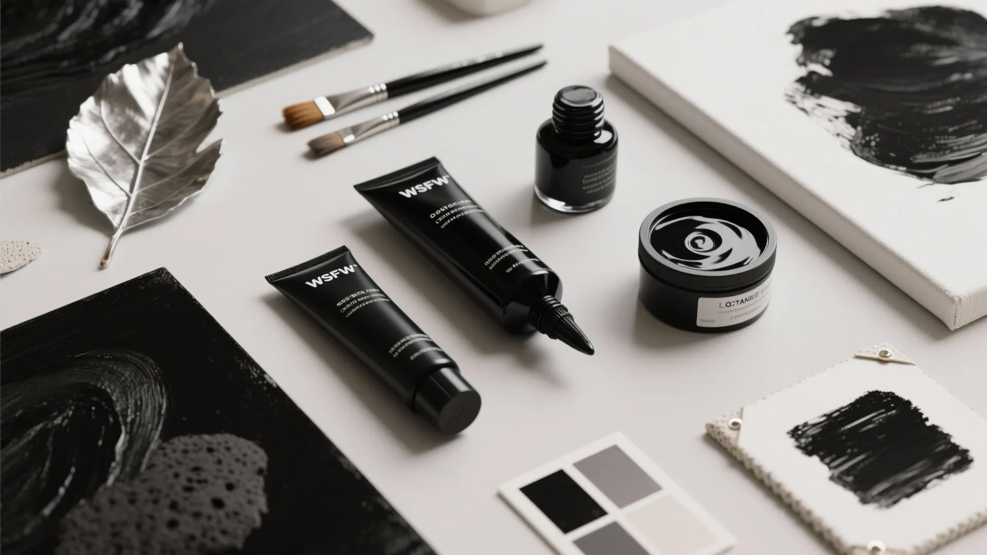 Flatlay of art materials: black pigments, acrylics, metallic leaf, brushes and small canvases.