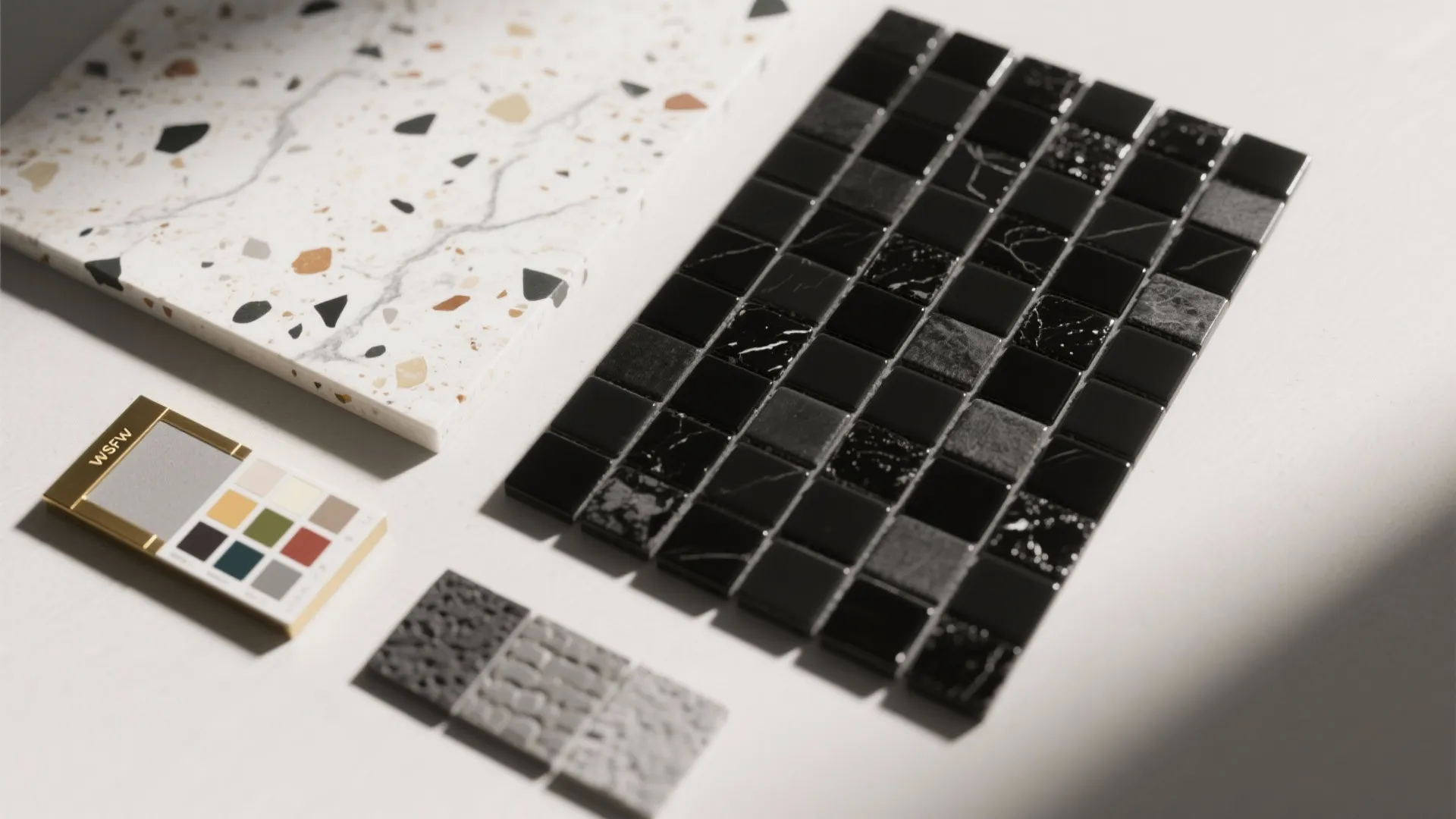 Flatlay of black mosaic tile, white marble slab and terrazzo sample with brass trim.