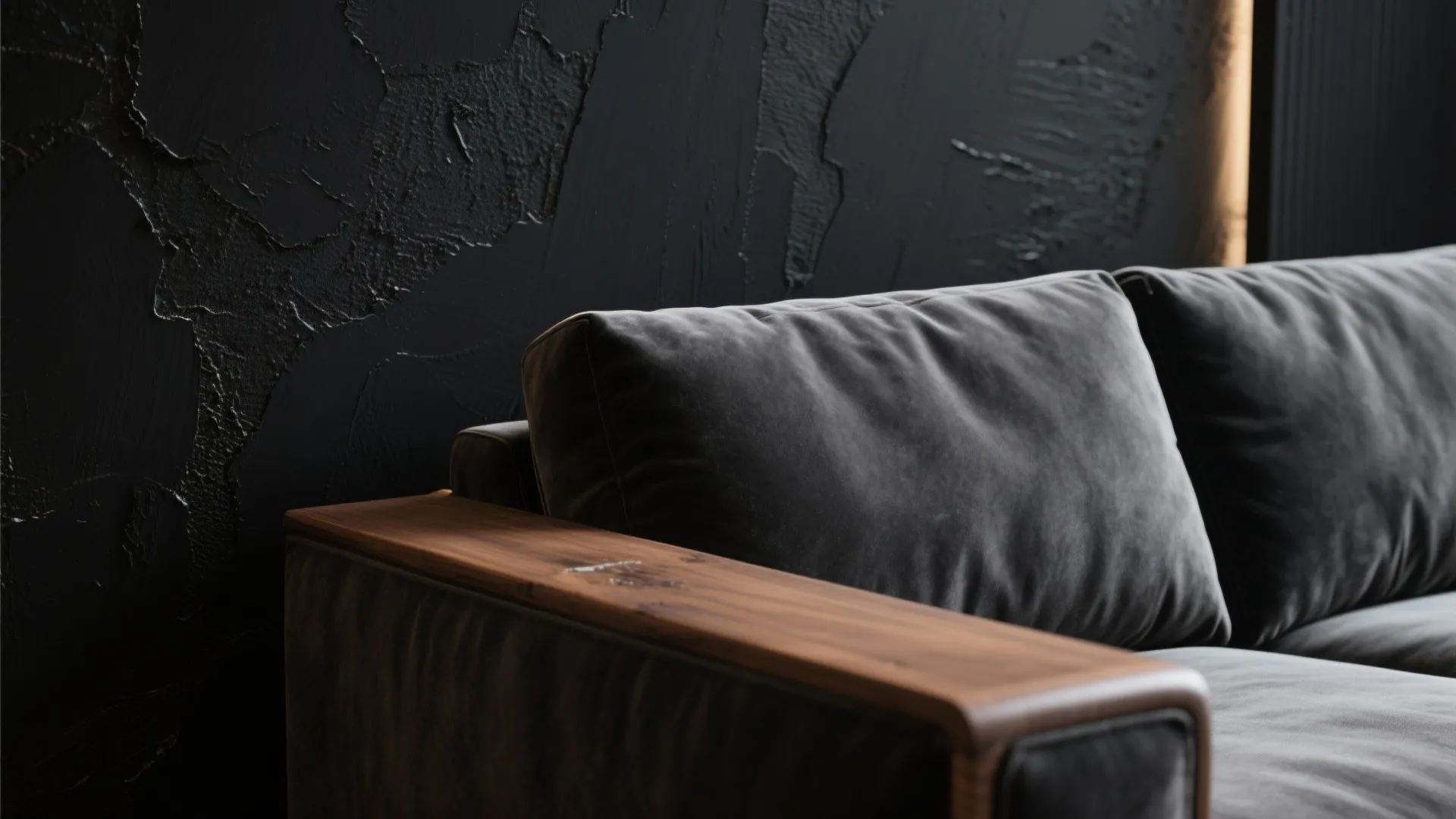 Close up of grey velvet sofa with wooden armrest against a textured matte black wall