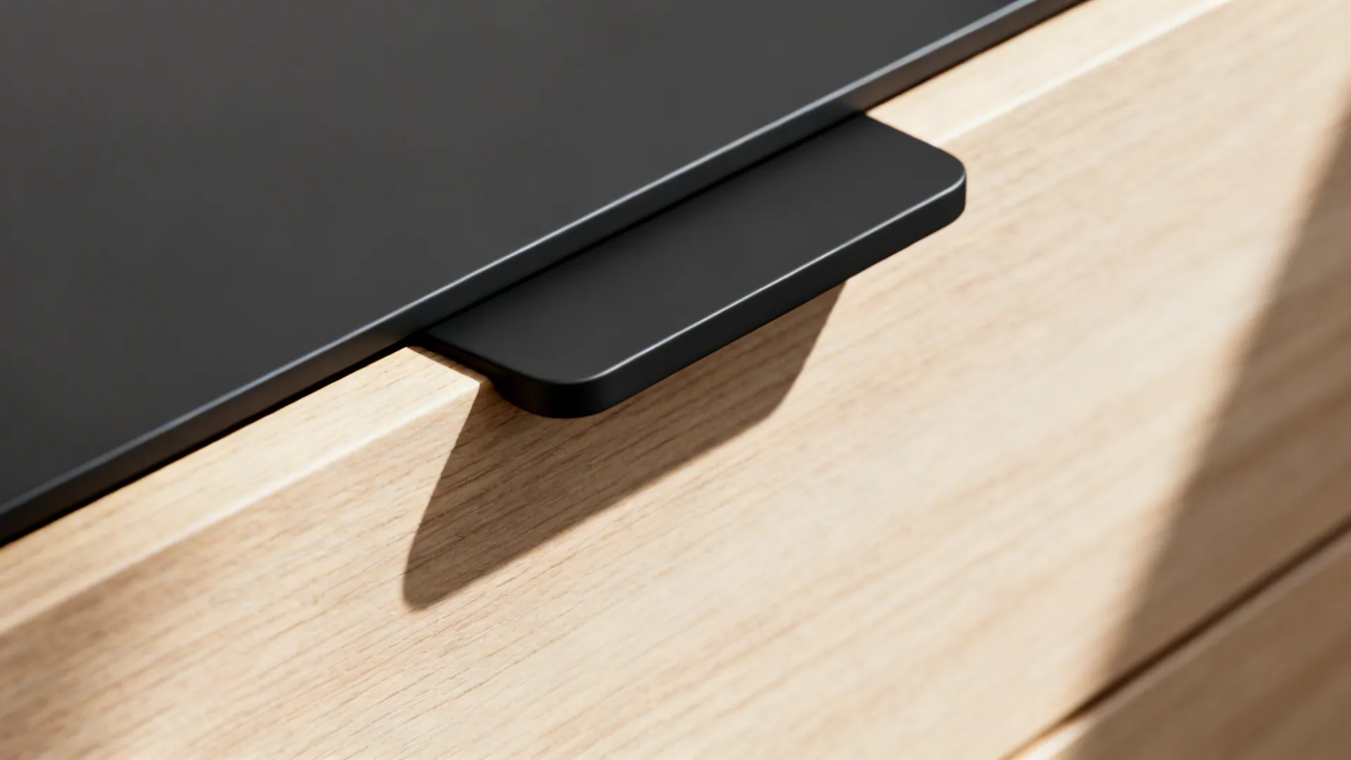 Macro of a matte black edge tab pull on a light wood slab drawer under soft daylight.
