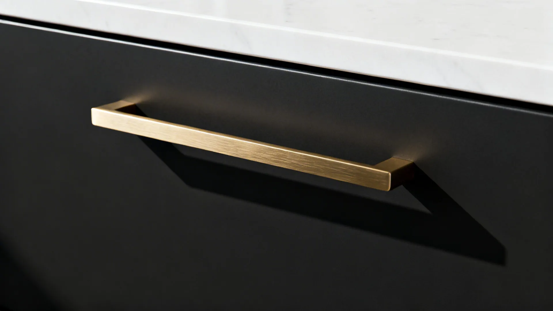 Macro detail of matte black cabinet with brushed brass pull and thin white quartz countertop.