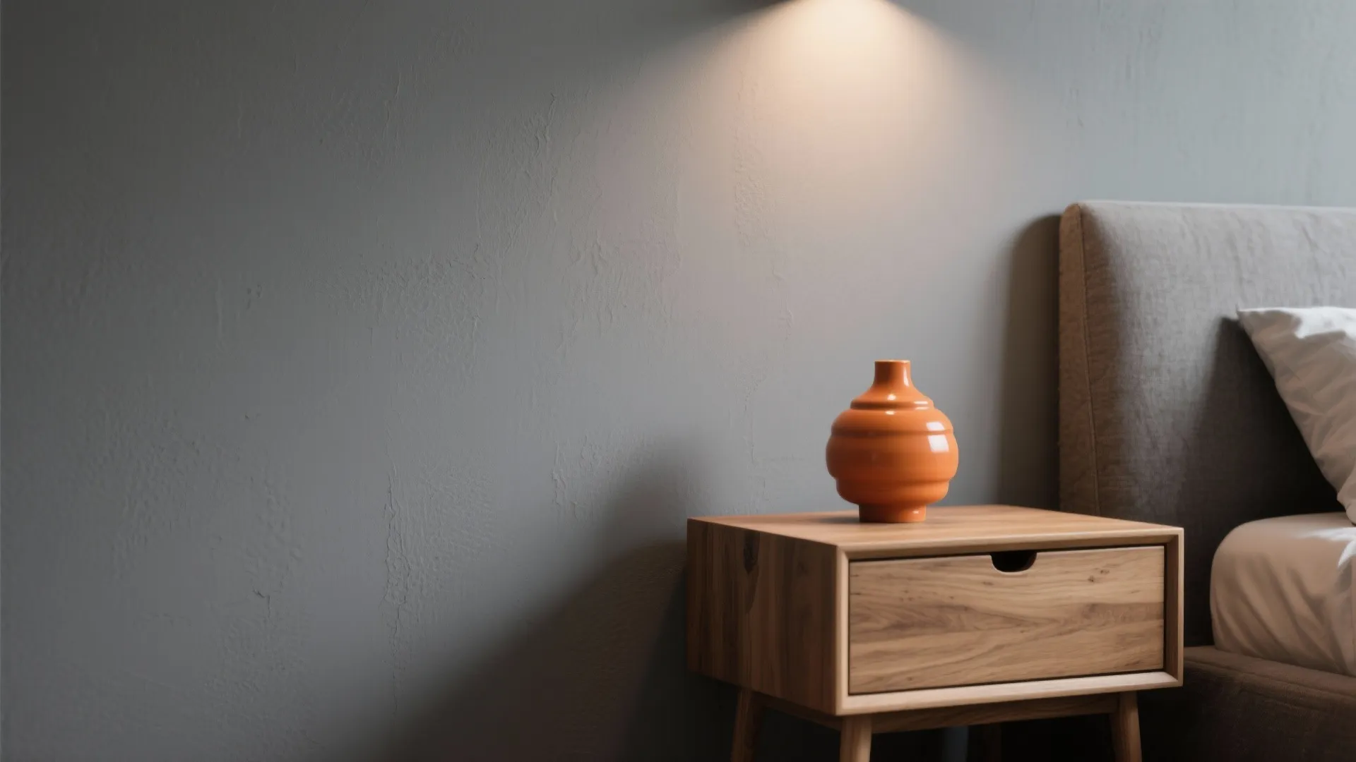 Modern bedroom with grey wall wooden bedside table orange vase and light from a wall light