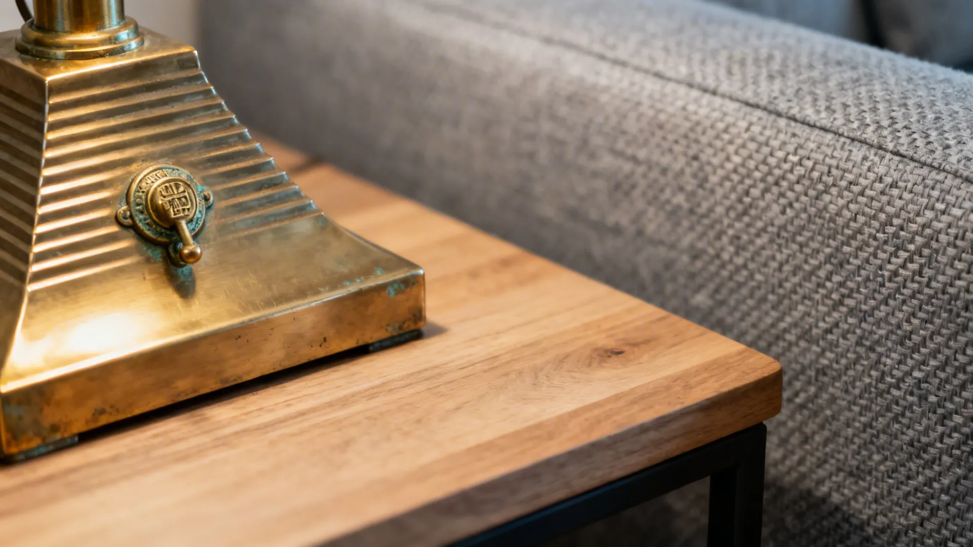 Brass lamp base and aged-gold hardware on a light oak table next to a grey sofa