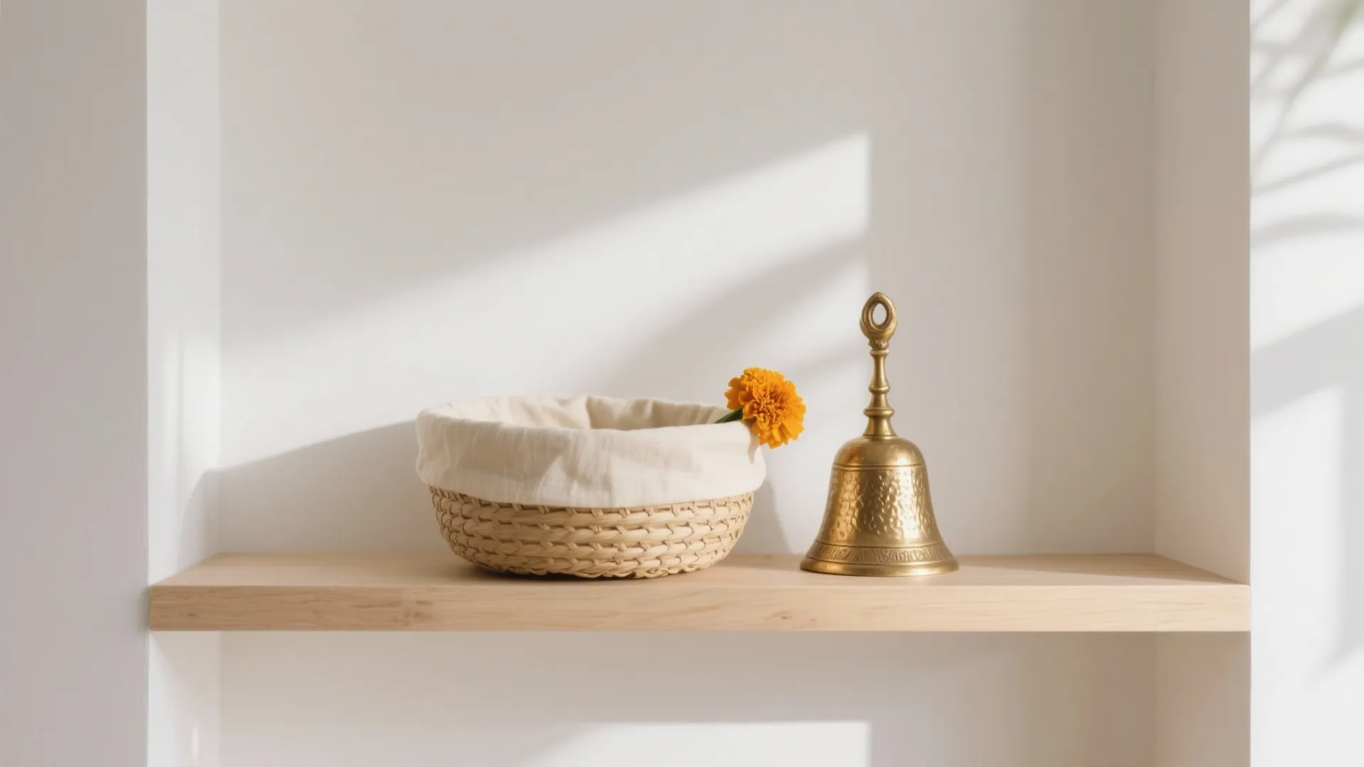 Minimalist natural-fiber pooja koodai with a brass bell and single marigold on a light wood shelf.
