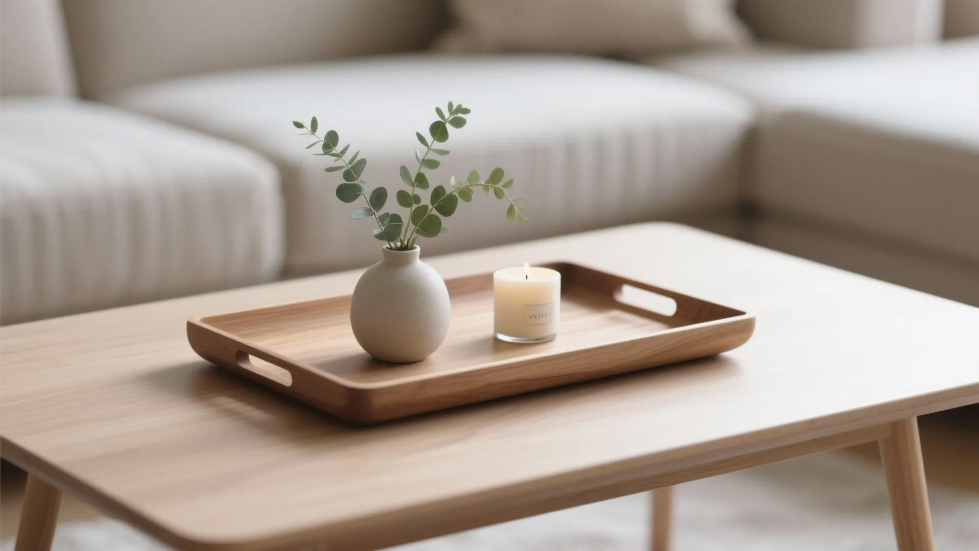 Wooden tray on coffee table holding small white vase with green leaves and lit candle