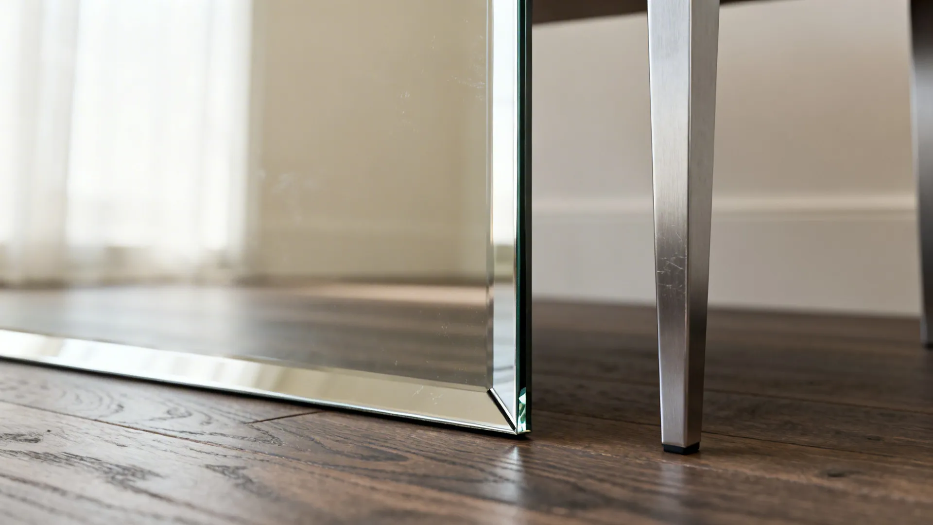 Macro of a tall mirror edge reflecting soft light beside a slim metal table leg on a wood floor.