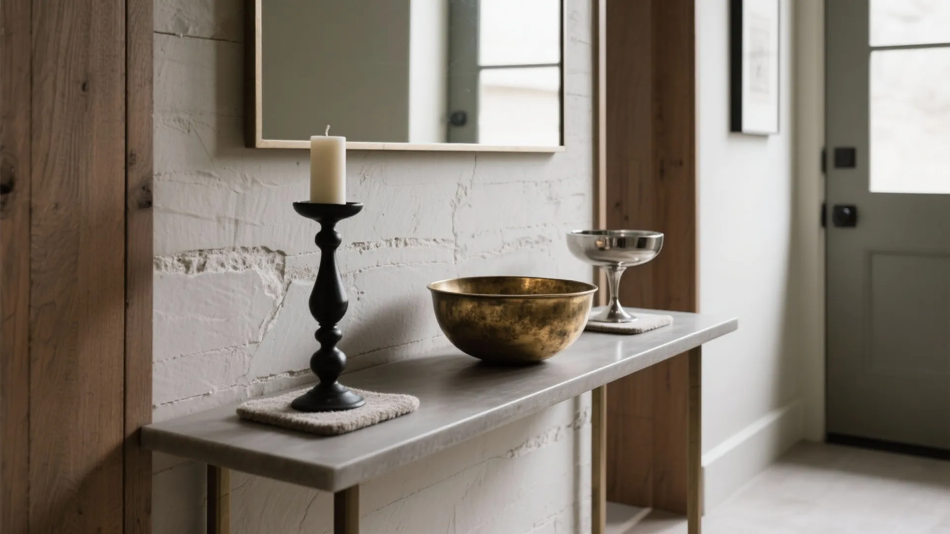 Slim console vignette with brass bowl, blackened steel candlestick, and nickel accent in a narrow entryway.