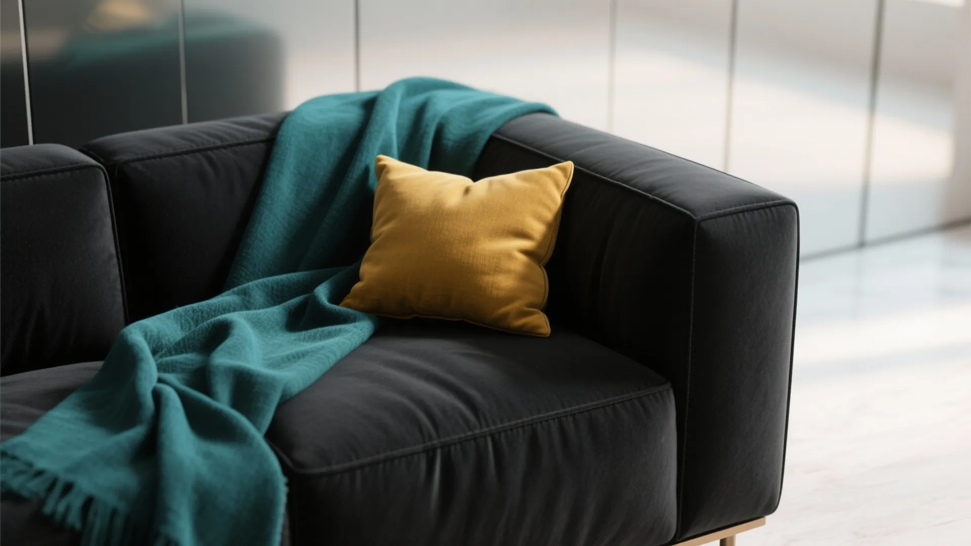 Close-up of a matte black sofa with a teal throw and mustard pillow against a light reflective wall.