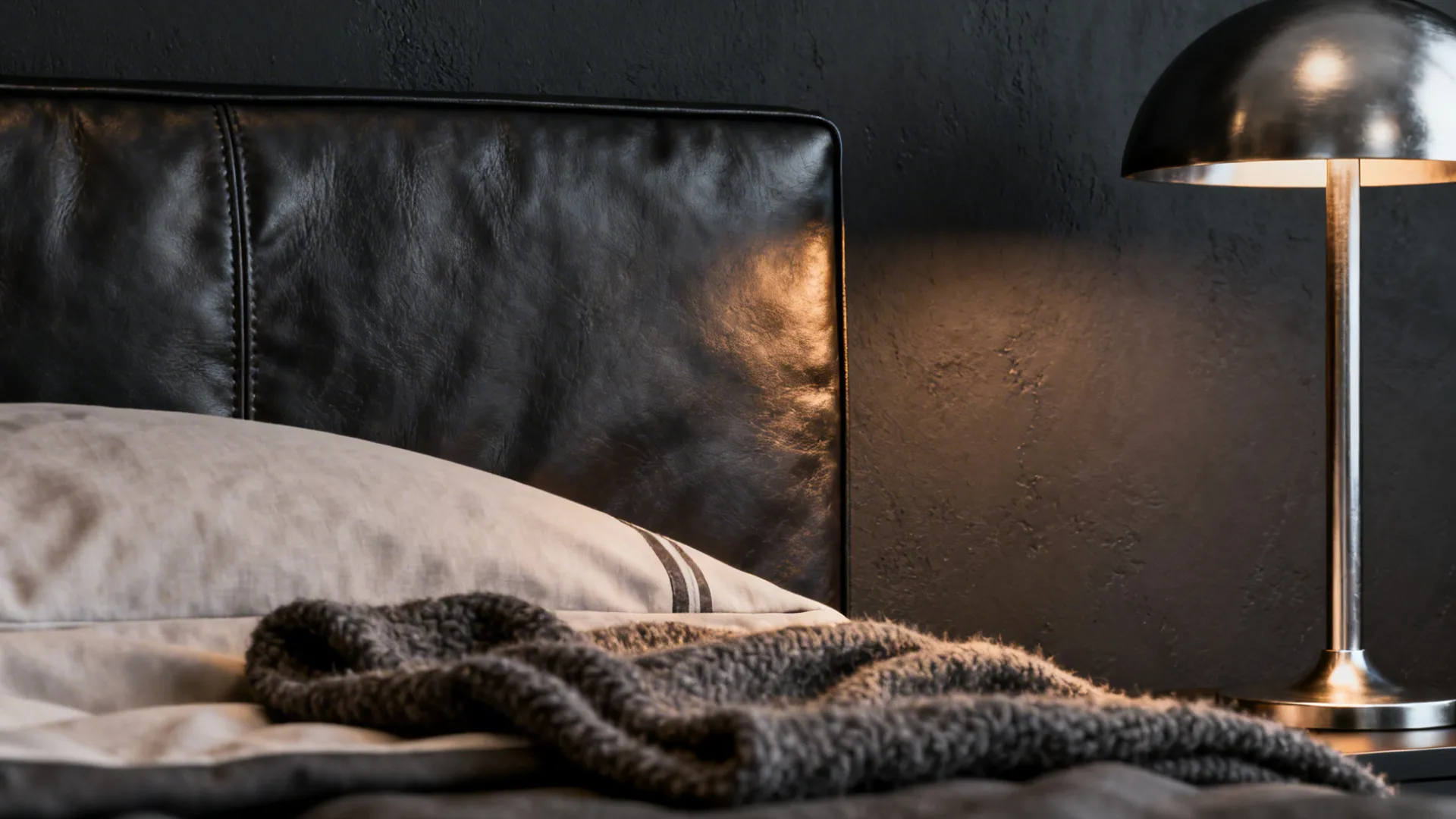 Black leather headboard with grey blanket and metal table lamp glowing against dark textured wall